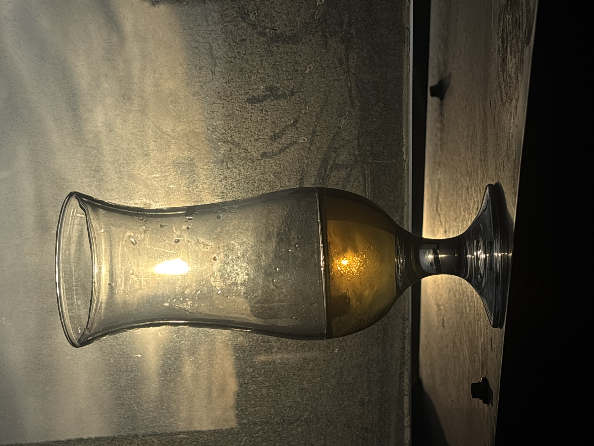 A glass vessel, possibly a stemmed beer glass, lies horizontally against a textured, possibly metallic or concrete, surface illuminated by a strong light source. The glass appears partially filled with a yellowish liquid. The stem of the glass extends toward the right edge of the frame, transitioning to a dark background. There are no people visible. There are no indications in the image of a specific location in Belgrade, Serbia.