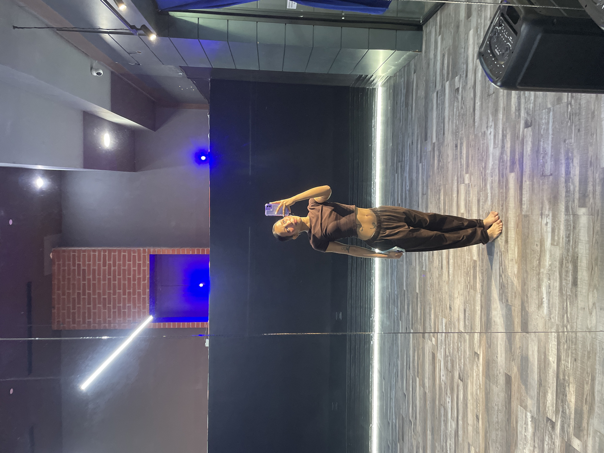 A full-body mirror selfie is taken by a person in a dance studio. The person is wearing a brown crop top and matching brown sweatpants, and is barefoot. The studio has a wooden floor and black walls, with a brick accent wall in the background. There are bright lights on the ceiling and a blue light emanating from an alcove. A large speaker is visible in the lower right corner of the image. The image is taken in Pampatar, Venezuela.