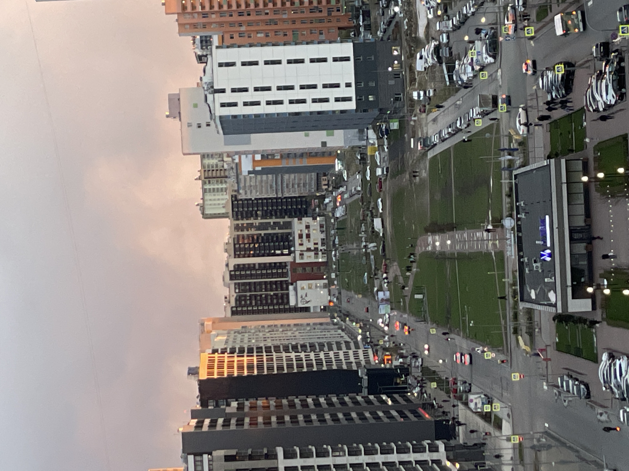 The image presents a high-angle view of a modern urban residential area, likely taken from a tall building. The original image is rotated 90 degrees counter-clockwise, making the buildings appear to lie on their sides.
The scene is dominated by numerous multi-story residential buildings, varying in height and design. Their facades feature a mix of colors, including white, grey, black, and some sections with orange or brown accents. Many windows are visible across these structures.
Several paved roads wind through the area, lined with a significant number of parked cars, mostly modern models in dark and light shades. Other vehicles are seen driving, indicated by illuminated headlights or taillights. Interspersed among the buildings and roads are patches of green grass, suggesting lawns or small park areas. Streetlights are visible along the roads and are illuminated, along with traffic lights.
The sky is overcast with a soft, muted light, suggesting either dawn, dusk, or a heavily clouded day. The illuminated streetlights further support the idea of twilight hours. A subtle warm glow can be observed in the sky, possibly hinting at a distant sunrise or sunset through the clouds.
A notable detail is a white sign with a blue "X" logo on a building in the lower-right portion of the image (when correctly oriented), likely indicating a commercial establishment. No other legible text is visible. The scene captures the functional landscape of a contemporary residential district in Murino, Russia, during what appears to be a calm transitional period of the day.
