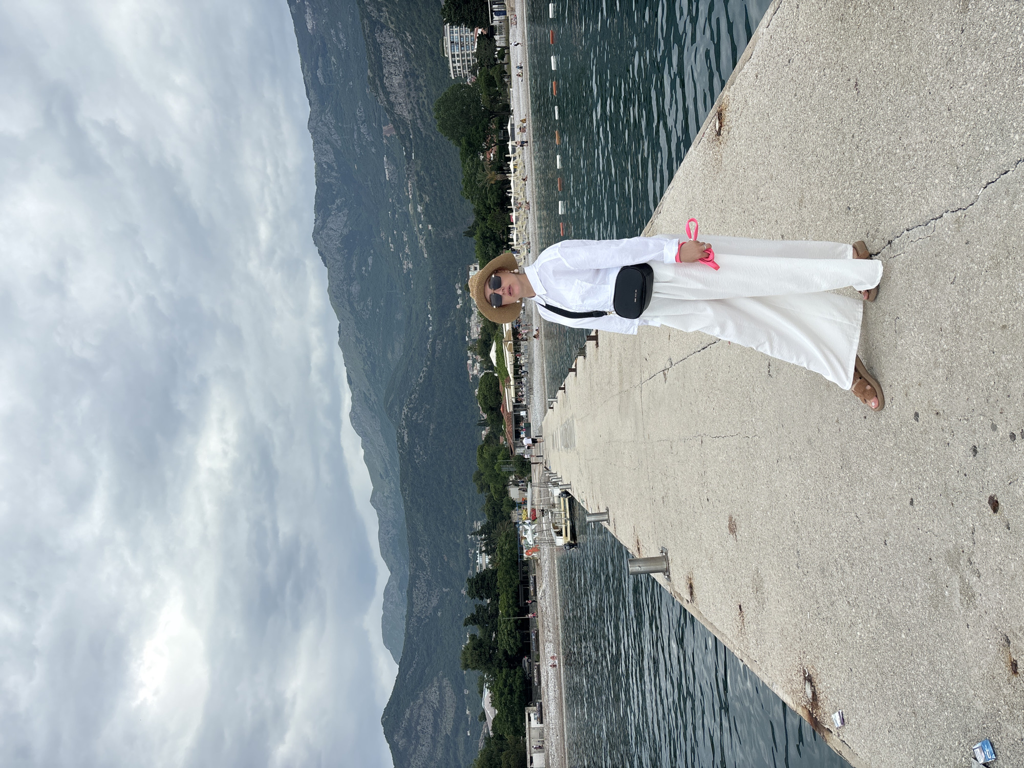 A woman is standing on the pier in Budva, Montenegro. She is wearing a white shirt and white pants and a straw hat, along with a black bag, and brown sandals. The background includes mountains, a beach, and the sea. The sky is cloudy. The pier is made of concrete and has metal poles.