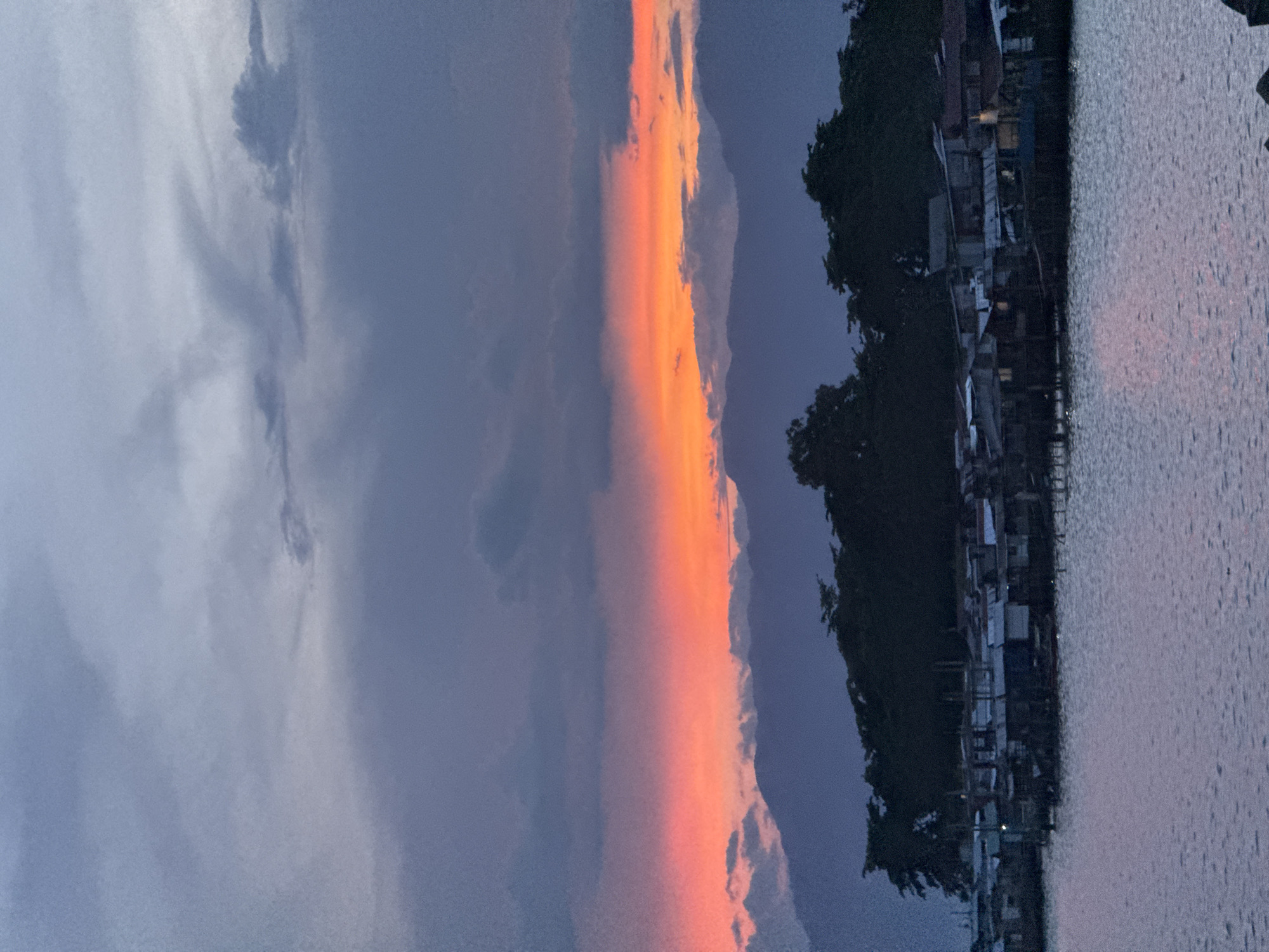 A landscape photograph taken in Davao City, Philippines, showing a body of water in the foreground and a coastline in the background. Along the coastline, there is a dense cluster of buildings and trees. The sky is filled with dark clouds on the left side of the image, and a brilliant orange sunset is taking place, filling up the central space and reflecting on the right side of the water. The image is taken vertically.
