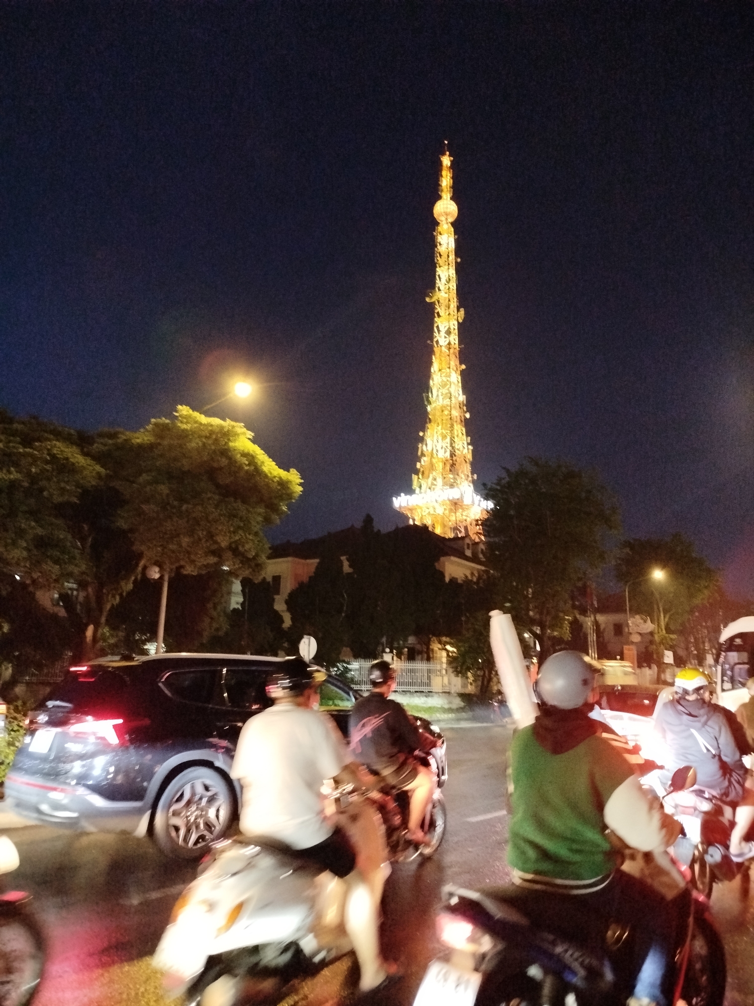 The image shows a nighttime street scene in Da Lat, Vietnam. The most prominent feature is the illuminated Da Lat TV Tower, a tall, latticed structure lit in yellow and gold, set against a dark blue sky. In the foreground, there is a street with a mix of traffic. Several motorbikes with riders are visible, some wearing helmets. A dark-colored SUV is partially visible on the left side of the frame. There are trees and streetlights lining the road, and buildings can be seen behind the trees. The atmosphere suggests an urban environment in the evening.