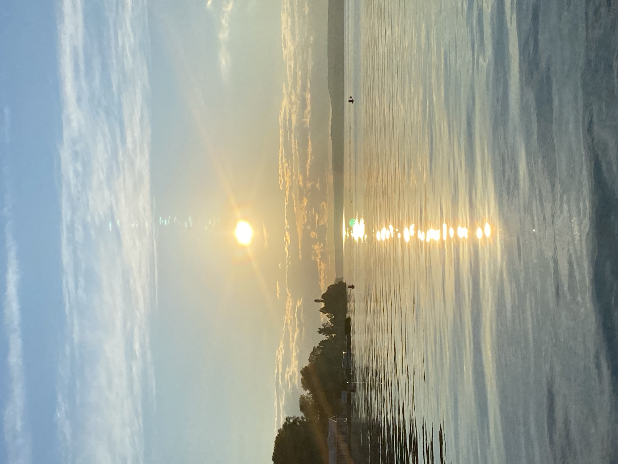 This is an image of a sunset over a body of water, likely a lake or river, on the shore of Votkinsk, Russia. The sun is low on the horizon, casting a golden glow across the sky and reflecting brightly on the water's surface. Wispy clouds are scattered across the blue sky. Along the left side of the image, there is a tree-covered shoreline with some structures visible, possibly houses or boathouses. A red buoy is visible in the water in the middle right of the frame, and another smaller dark object is visible further out. The water exhibits gentle ripples, creating a shimmering effect from the sunlight. The scene is tranquil and serene, depicting a natural landscape at dusk.