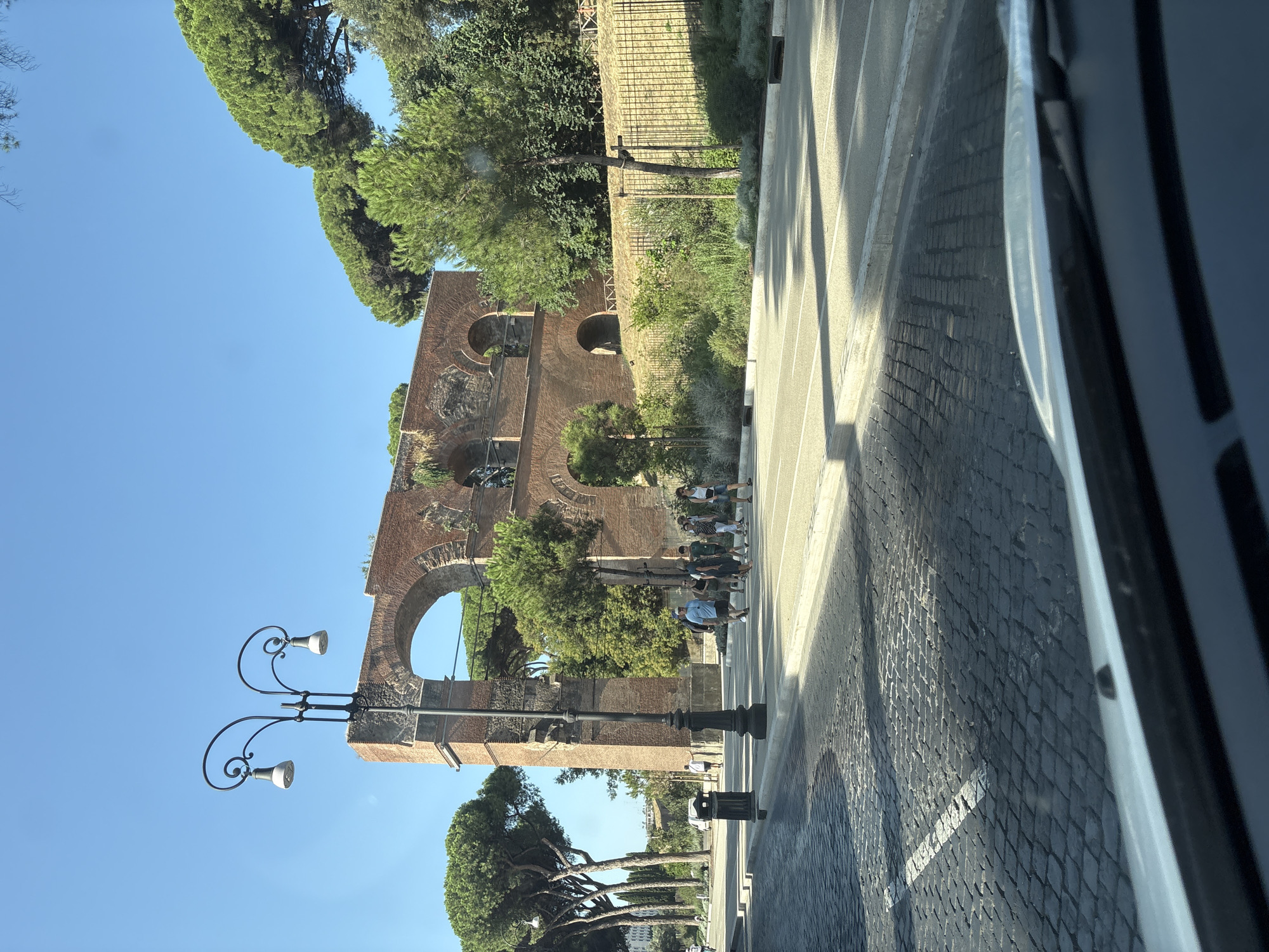 The image shows an exterior view of the Aqua Claudia, an ancient Roman aqueduct, in Rome, Italy. The aqueduct is constructed of red brick and features multiple arches. In the foreground, a street with cobblestone paving leads to a pedestrian crossing. A lamppost with two ornate fixtures stands to the left of the aqueduct. A group of people are walking across the pedestrian crossing. Behind the aqueduct, lush green trees and foliage are visible, with a clear blue sky overhead. The scene appears to be captured on a sunny day.