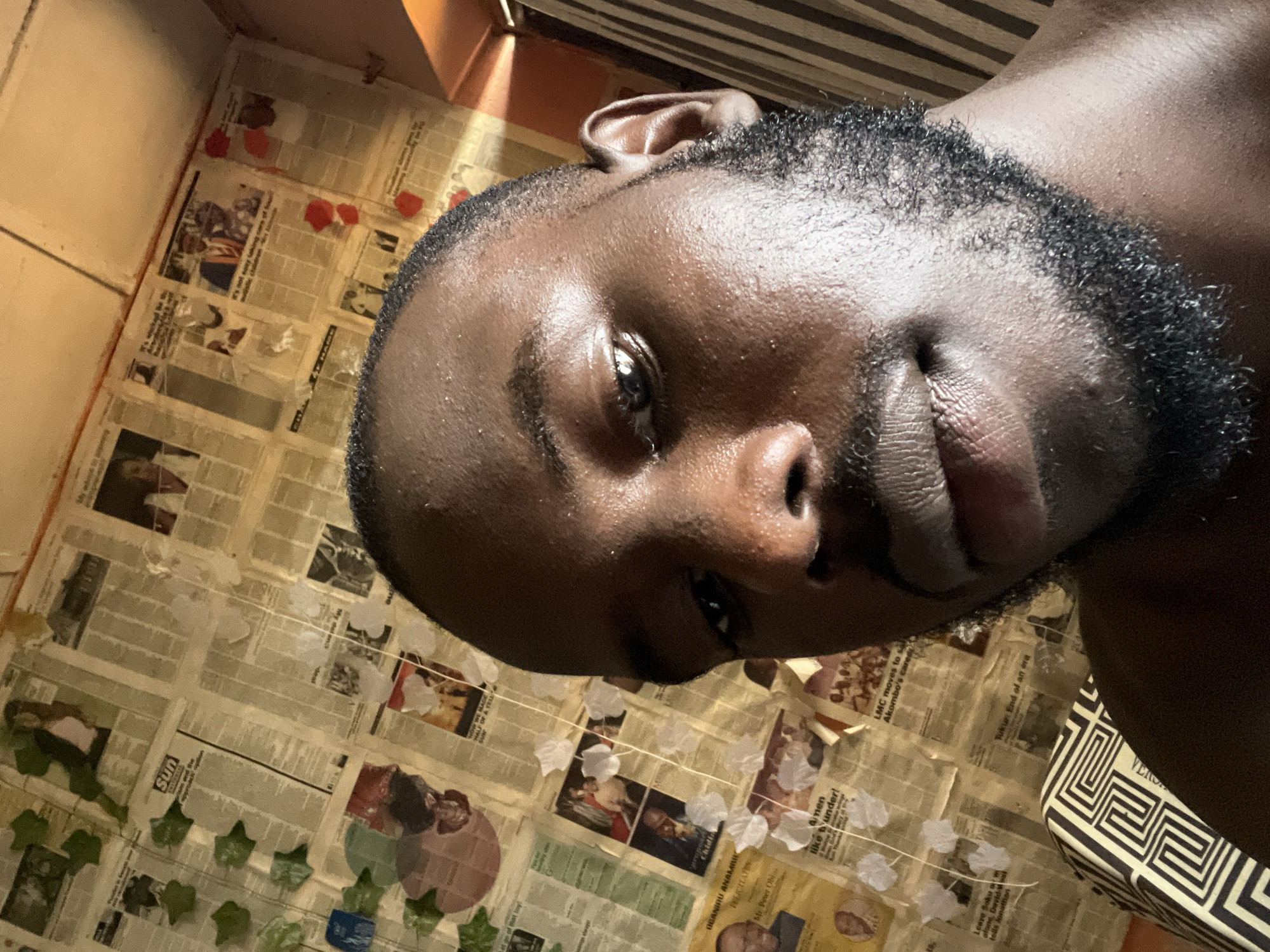 This is a close-up, low-angle selfie of a young Black man with dark skin, a beard, and short hair. He is looking directly at the camera, with his head tilted to the side. His eyes are open, and his mouth is slightly parted, conveying a relaxed or contemplative expression. He is partially illuminated by a shaft of sunlight coming from the left, highlighting the texture of his skin.
The background appears to be a wall covered in newspaper clippings, with some decorative elements like artificial green leaves and white paper flowers. The newspaper articles are partially visible, and some text is legible, including headlines and names. The overall setting seems to be indoors, possibly in a room decorated with these newspaper clippings. The orientation of the image suggests it was taken in Rafin Par, Nigeria, as per the location context. The time of day is likely daytime, given the sunlight.