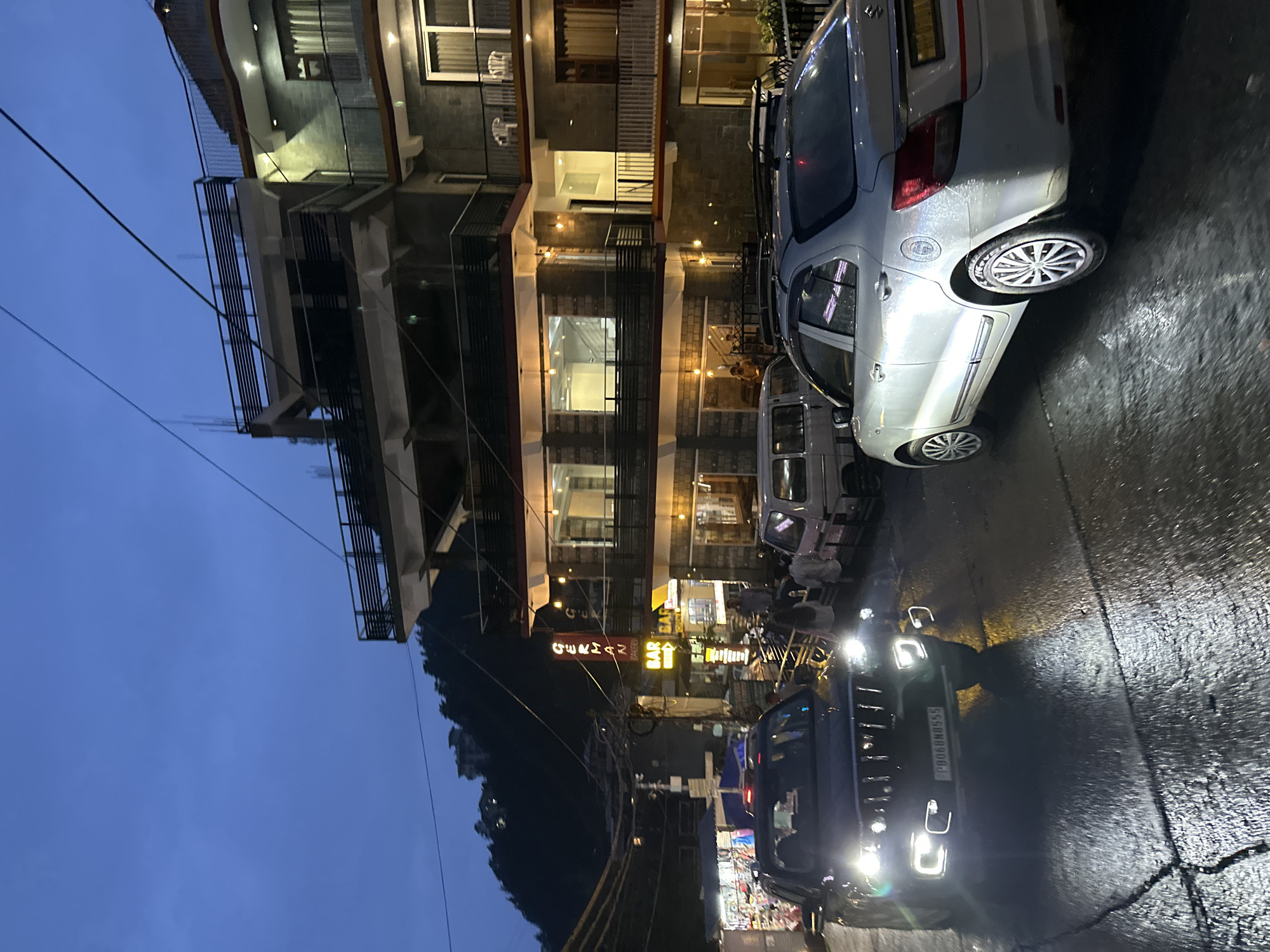 The image depicts a street scene at dusk or night in a town with hilly terrain. A multi-story building, illuminated by warm interior lights and exterior lamps, dominates the right side of the frame. The building features balconies with railings and large windows. In front of the building, a street is wet, reflecting the lights. A dark-colored SUV with its headlights on is positioned in the foreground on the left, facing towards the viewer. To its right, a white sedan is parked, also illuminated by streetlights. Several people are visible on the sidewalk and near the vehicles, appearing to be pedestrians or shoppers. Signs for "GERMAN BAKERY" and "BAR" are visible on the building, indicating commercial establishments. Power lines crisscross the sky above the street. The overall atmosphere suggests a bustling urban or tourist area at twilight.