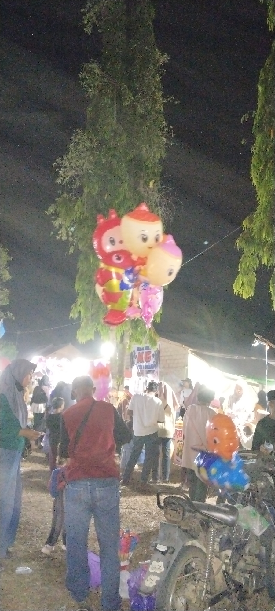The image captures a night scene in the city of Sido Rahayu, Indonesia, likely during a local event or market. In the foreground, a person wearing a red shirt and blue jeans is seen from the back, standing on a dirt or grass ground. Next to them, to the right, is the rear of a motorcycle, partially visible and appearing dusty. Several colorful balloons, including cartoon character shapes like a red devil-like figure and characters with round faces, are clustered together and appear to be floating. In the background, a crowd of people are milling around, with some standing near stalls or tents illuminated by artificial lights. The lighting suggests it is nighttime, and trees with dense foliage are visible on the left and right sides of the frame, framing the scene.