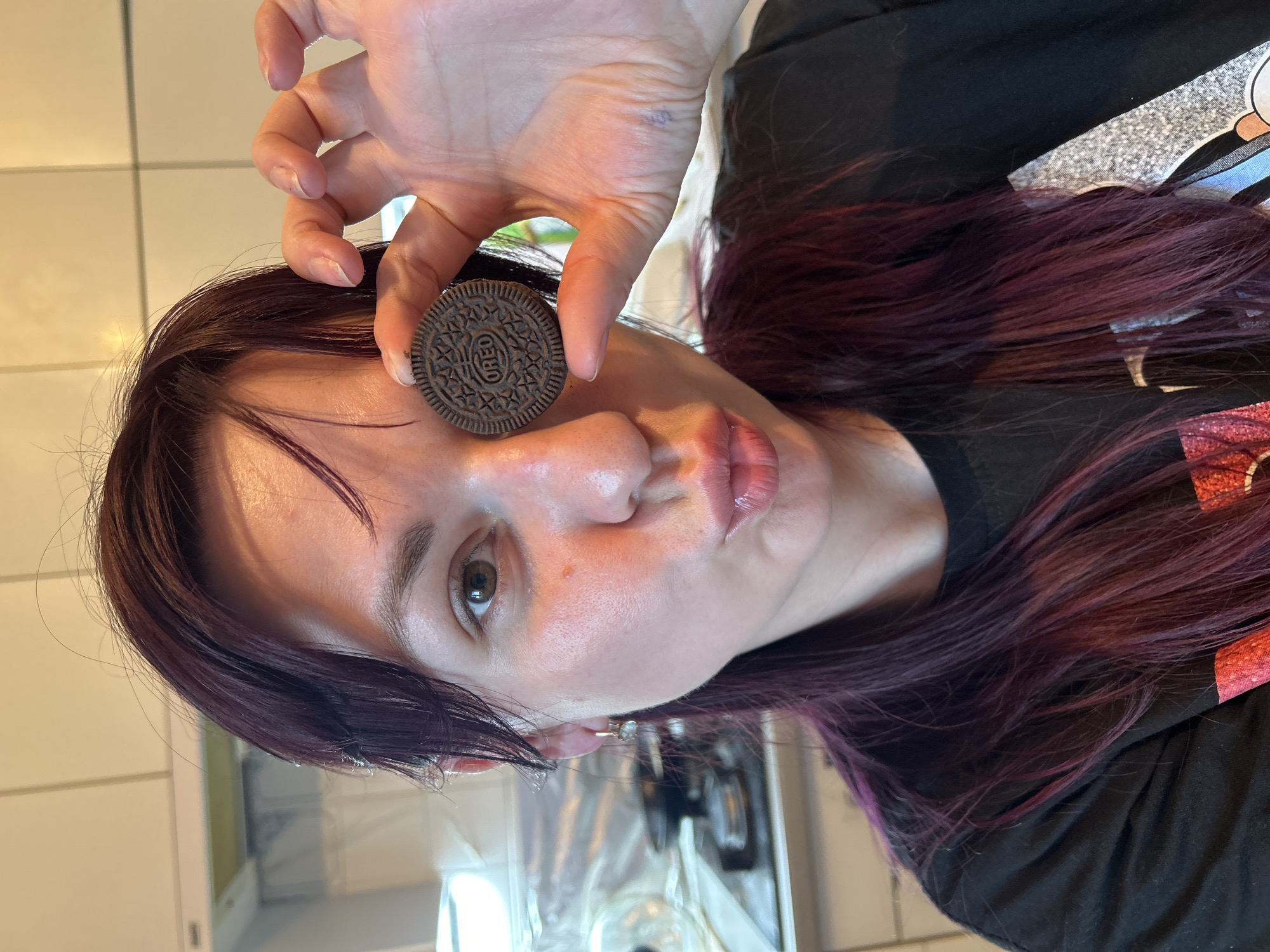 A woman with dark, purple-tinted hair holds a chocolate cookie with a white cream filling, possibly an Oreo, up to her right eye. She is making a kissing face, with her lips pursed. She is wearing a black t-shirt with a graphic design on it. The background appears to be a kitchen, with white tiled walls and a stovetop visible. The image was taken in the city of Фоки, Russia.