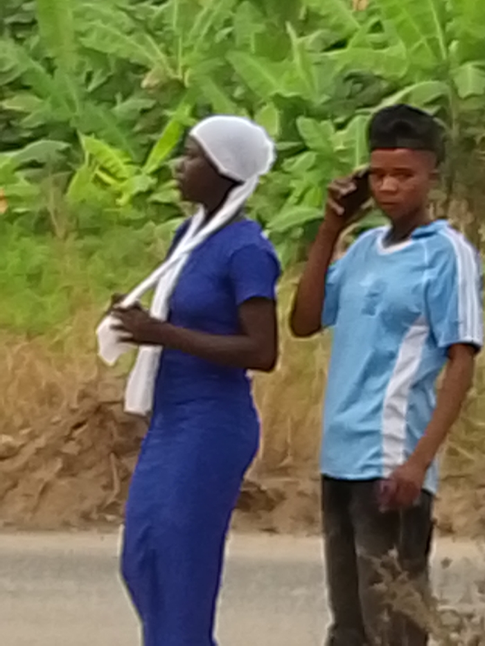 The image shows two young individuals standing outdoors during what appears to be daytime. The background is dominated by lush green foliage, possibly banana plants or similar large-leafed vegetation, suggesting a tropical or subtropical environment, or a rural/semi-urban setting.
On the left, a person, likely a young woman, is visible in profile facing left. She has dark skin and is wearing a white head covering or wrap, part of which hangs down over her shoulder and is held in her hands. She is dressed in a long-sleeved, royal blue dress that extends down her body. Her posture is relaxed, with her head slightly tilted up.
On the right, another person, likely a young man, is partially visible, facing slightly towards the left side of the frame but looking forward. He also has dark skin and is holding a dark-colored smartphone to his right ear, seemingly in a call. He has short, dark hair with a distinct haircut. He is wearing a light blue short-sleeved t-shirt with white stripes or panels on the sides and sleeves. The bottom part of his attire consists of dark pants.
The ground beneath them appears to be an unpaved road or dirt path, with some brown earthy tones visible. The overall image quality is somewhat soft or blurry, but the key details of the individuals and their actions are discernible. There is no visible text in the image.