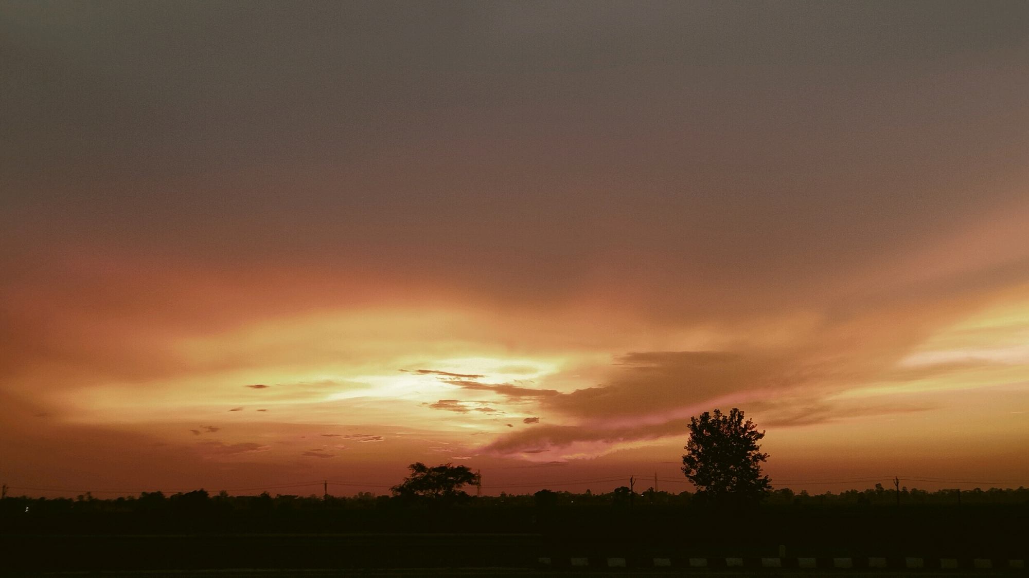 The image depicts a sunset over a rural landscape in Miranpur, India. The sky is filled with dramatic clouds painted in hues of orange, yellow, and pink, with a gradient of darker tones towards the top. Silhouetted against the vibrant sky is a flat horizon line of trees and vegetation. In the foreground, a dark silhouette of the ground is visible, suggesting a field or open area. A solitary, full-bodied tree stands prominently in the mid-ground on the right side of the frame, its branches reaching upwards. Faint outlines of utility poles and wires are also visible along the horizon, indicating the presence of infrastructure. The overall scene is serene and natural, capturing the beauty of a sunset in a rural Indian setting.