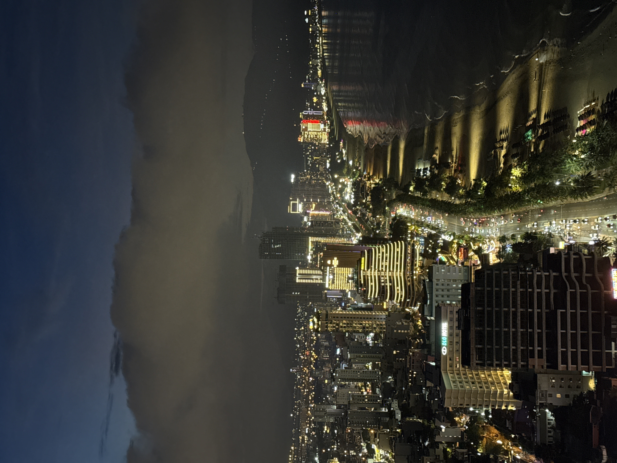 An aerial night view captures the city of Da Nang, Vietnam, showcasing its coastal urban landscape. The image is dominated by a dense city skyline with numerous illuminated skyscrapers and buildings. A prominent beach runs along the coastline, with palm trees and beach chairs visible. The ocean meets the beach, with gentle waves lapping the shore. A street lined with cars and streetlights runs parallel to the beach, connecting the city to the coast. In the background, a range of mountains is faintly visible against the twilight sky, which is partly obscured by clouds. The scene conveys the vibrant nightlife and coastal charm of Da Nang.