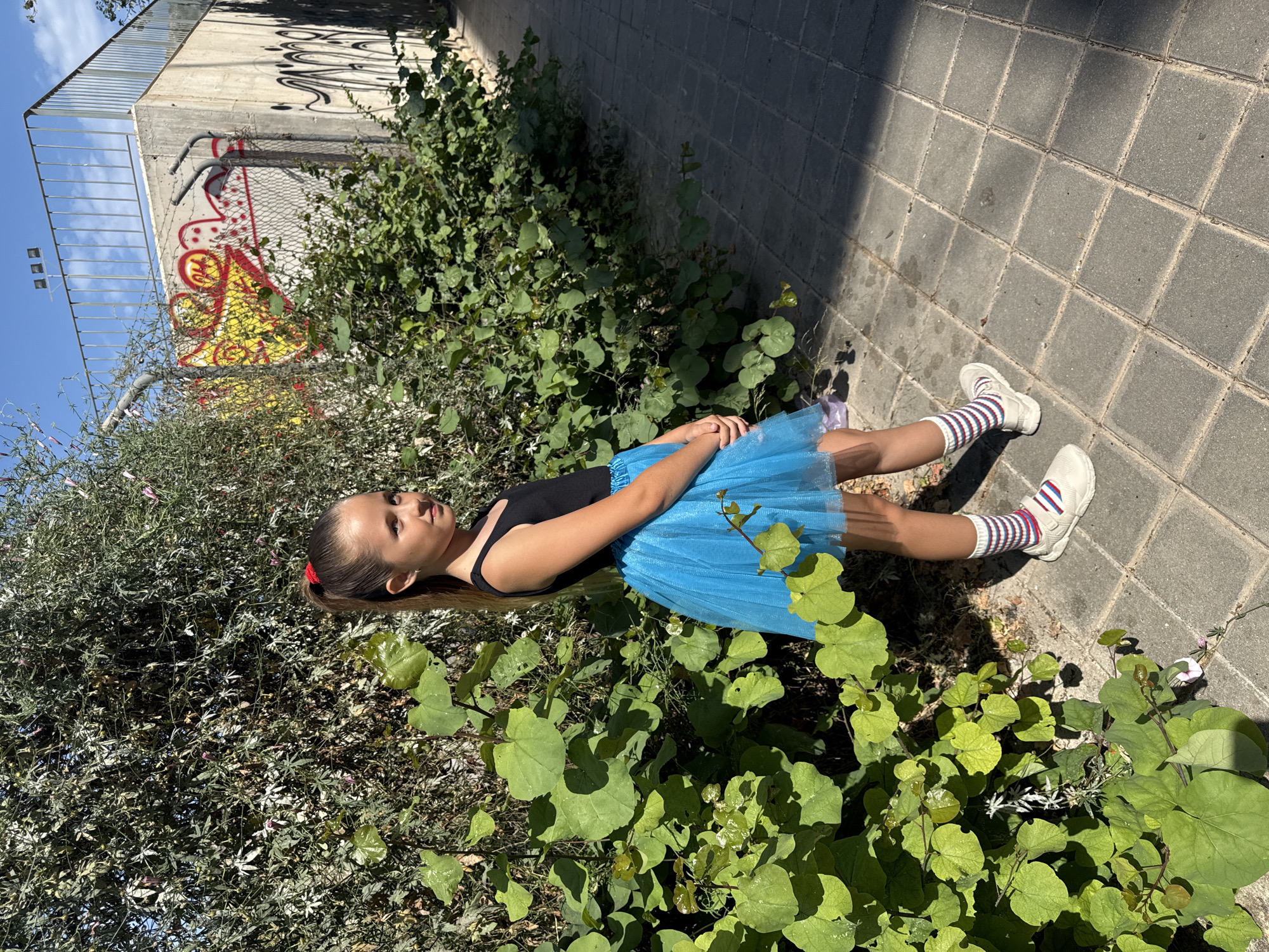 A young girl lies amongst dense vegetation on a tiled ground surface in an urban environment. She is dressed in a black leotard and a blue tulle skirt, and she wears sneakers with striped socks. Her hair is pulled back, with a red hair tie visible. The vegetation surrounds her, and a concrete wall with graffiti is seen in the background. The sky is visible in the upper left.
