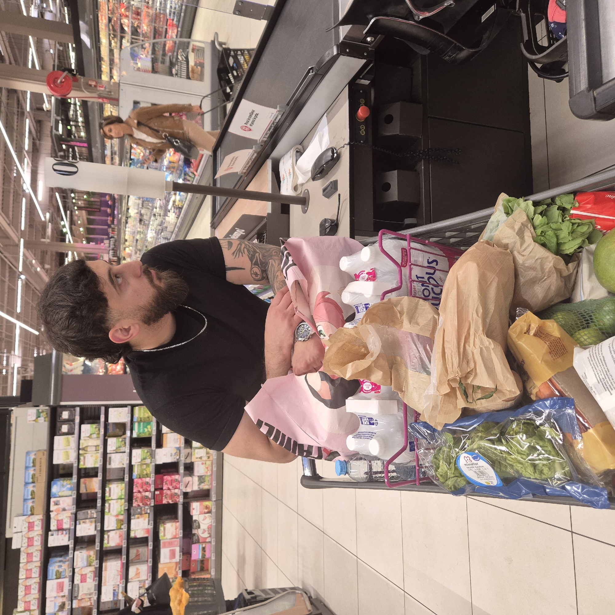 The image shows a man at a checkout counter in a supermarket. He has dark hair, a beard, and tattoos on his arm. He is wearing a black shirt and a silver chain. His shopping cart is full of groceries, including food items in brown paper bags, bottled water, and packaged lettuce. A pink Anime shopping bag is visible in the cart. The checkout counter is equipped with a scanner and other point-of-sale equipment. Aisles of shelved products are visible in the background, indicating the store's layout. A woman is seen in the background near a checkout line. The OCR text suggests the scene is in Geispolsheim, France, possibly at a shop like Mimafior.