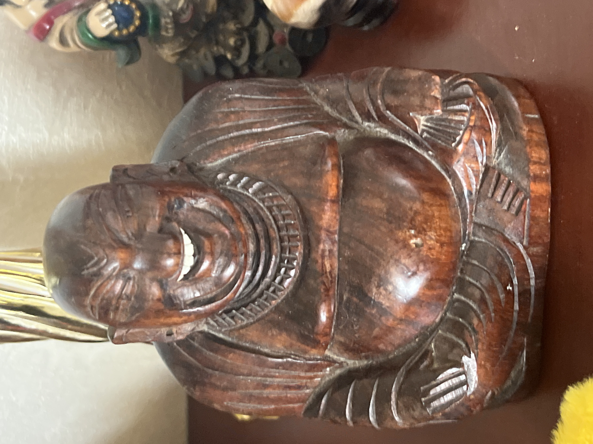 A close-up, eye-level shot shows a wooden statue of a laughing Buddha. The statue is dark brown with carved details. The Buddha is depicted with a large, smiling face, closed eyes, and a full belly. He is wearing a robe and a necklace. The statue is sitting on a dark brown surface, possibly a table or shelf. In the background, to the right of the Buddha, another statue of a figure in colorful clothing can be partially seen. To the left of the Buddha, a portion of a tall, golden, reflective object is visible. The lighting is soft, creating some reflections on the polished wood of the Buddha statue. The background is out of focus, with a light beige wall visible.