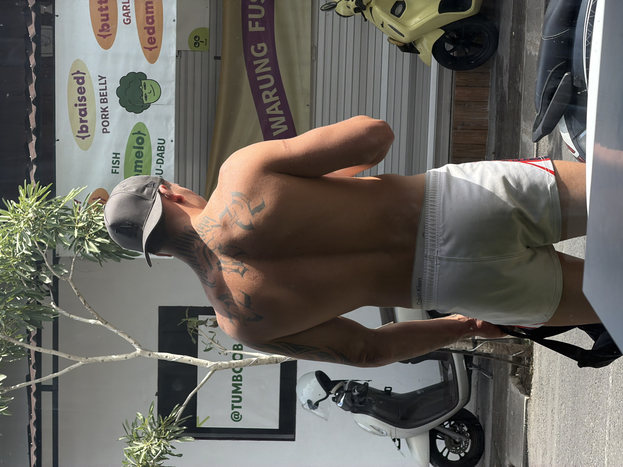 A shirtless man wearing white Calvin Klein boxer shorts and a grey baseball cap is standing with his back to the camera. He has extensive black tattoos on his back and arms. He is carrying a black bag in his left hand. In the background, a white scooter with a helmet on it is parked to the left, and a yellow scooter is parked to the right. A building with a striped metal facade and a banner reading "WARUNG FUS" is behind the scooters. Signs on the building include "@TUMBOOB" and an advertisement with text like "{braised} PORK BELLY", "FISH {melo} U-DABU", "{butter} GARLIC", and "{edamame}". The man is standing on a paved street in front of the building. The scene appears to be in the city of Mengwi, Indonesia.