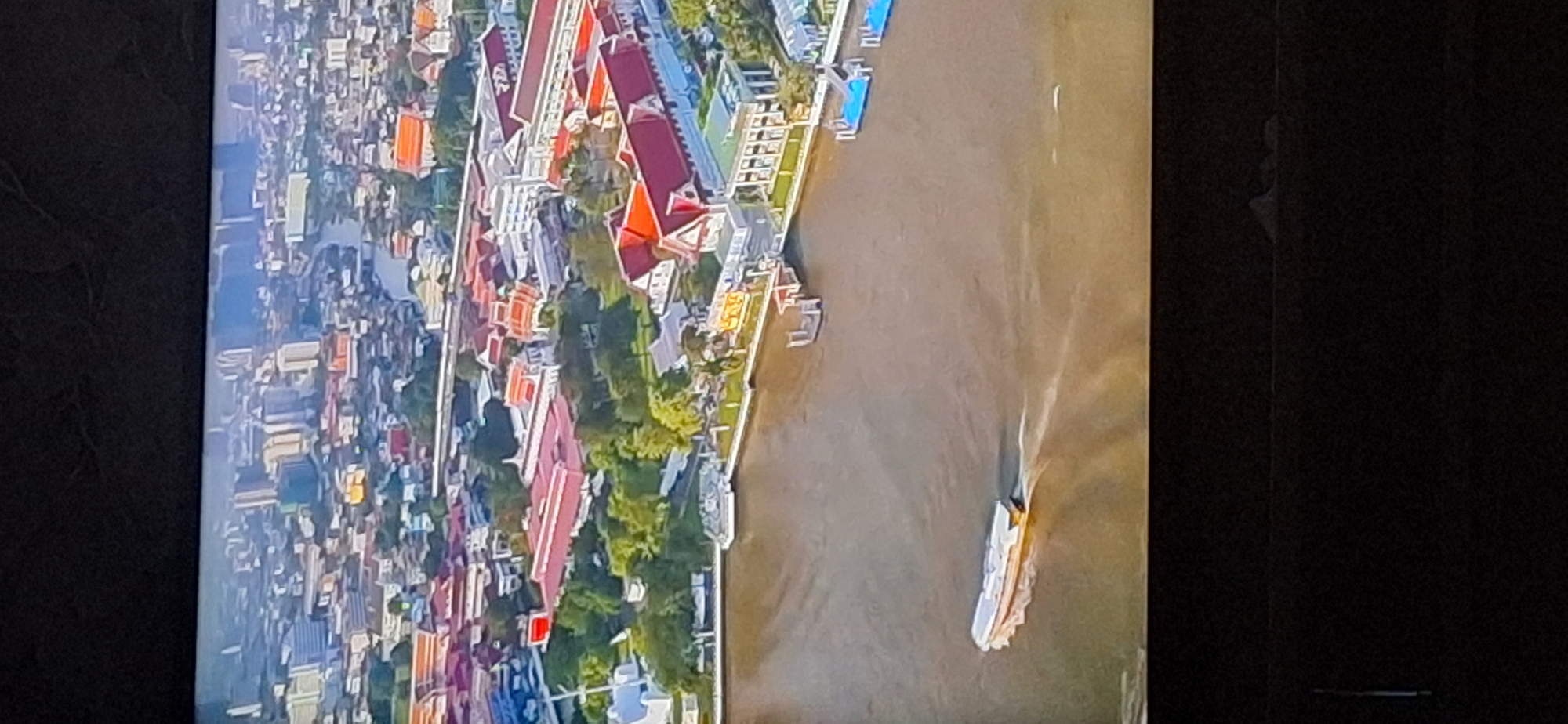 The image is an aerial view of a city with a river running through it. The city has a mix of low-rise buildings with red tile roofs and taller modern buildings in the background. Along the riverbank, there are several structures with ornate architecture and red tile roofs, suggesting they might be historical or religious sites. A long, narrow boat is moving on the murky brown water of the river, leaving a wake behind it. The overall scene depicts a bustling urban environment situated alongside a significant waterway, with a blend of traditional and contemporary architectural styles. There is no indication of the city being Bila Tserkva, Ukraine in the provided image.