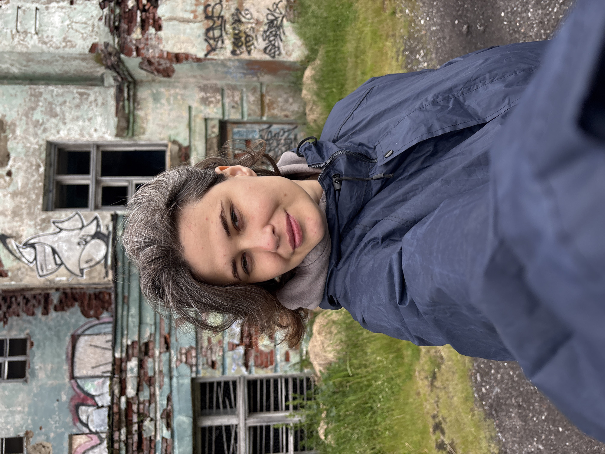 A selfie taken by a woman with graying brown hair, wearing a dark blue hooded jacket and a grey hoodie, is presented. She is positioned in the foreground, slightly to the right of center, facing the camera with a gentle smile. Her hair is swept back from her face, revealing her forehead and cheeks. Behind her, an abandoned, dilapidated building stands prominently. The building's facade is weathered, with peeling blue paint revealing red brick beneath. Graffiti, predominantly in black and white, adorns the walls and windows of the structure. Several windows are boarded up or broken, with one larger window on the right side of the frame showing a dark, empty interior. Overgrown green grass and scattered rocks surround the building, suggesting a neglected outdoor environment. The overall scene evokes a sense of decay and abandonment in a rural or semi-urban setting, possibly within the city of Teriberka, Russia, known for its abandoned structures.