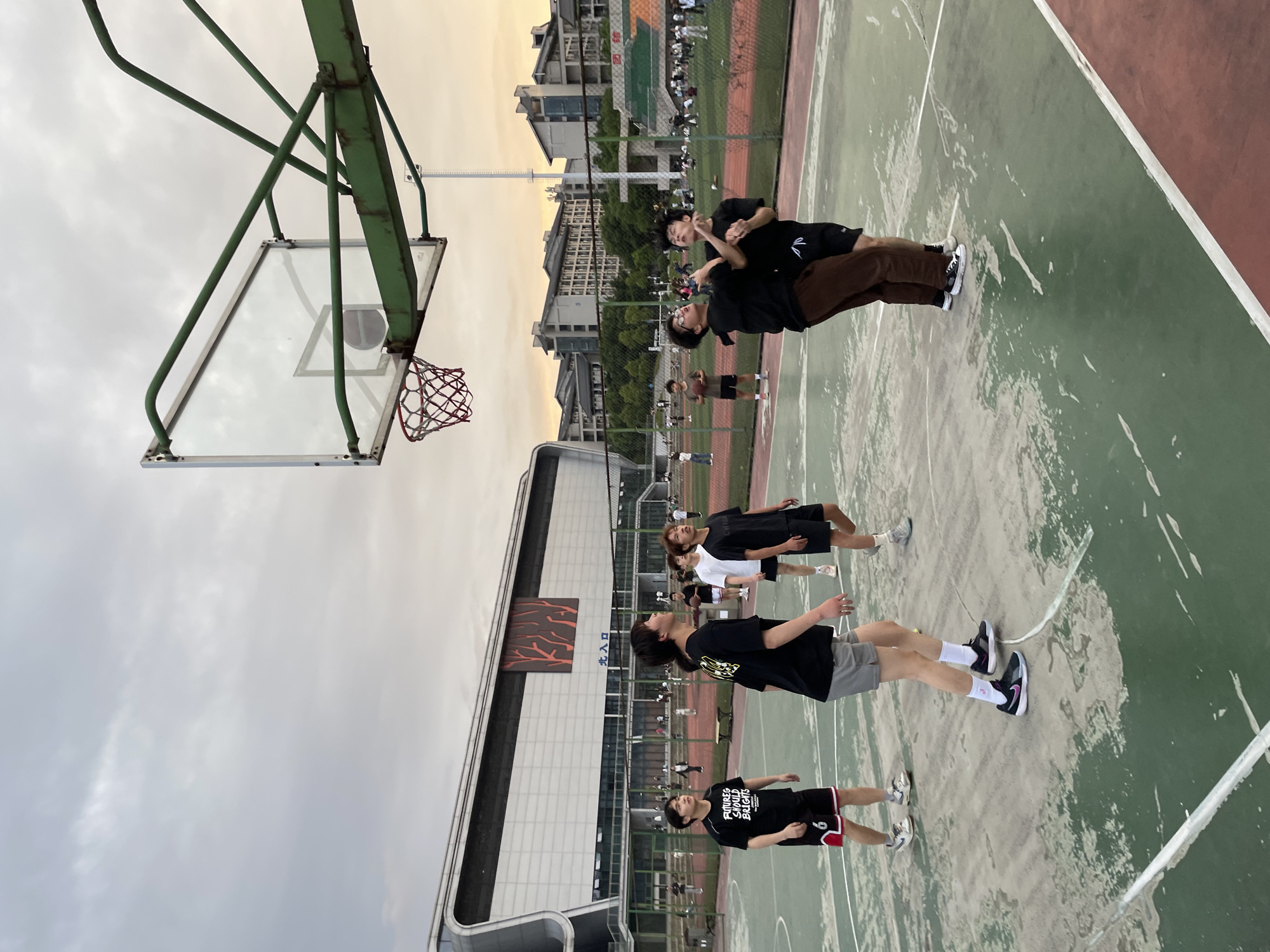 The image shows a group of five people standing on a basketball court in Jiangjin, China. The court is marked with painted lines and has a basketball hoop on one end. The backdrop features buildings and a sky with clouds. People appear to be interacting casually.