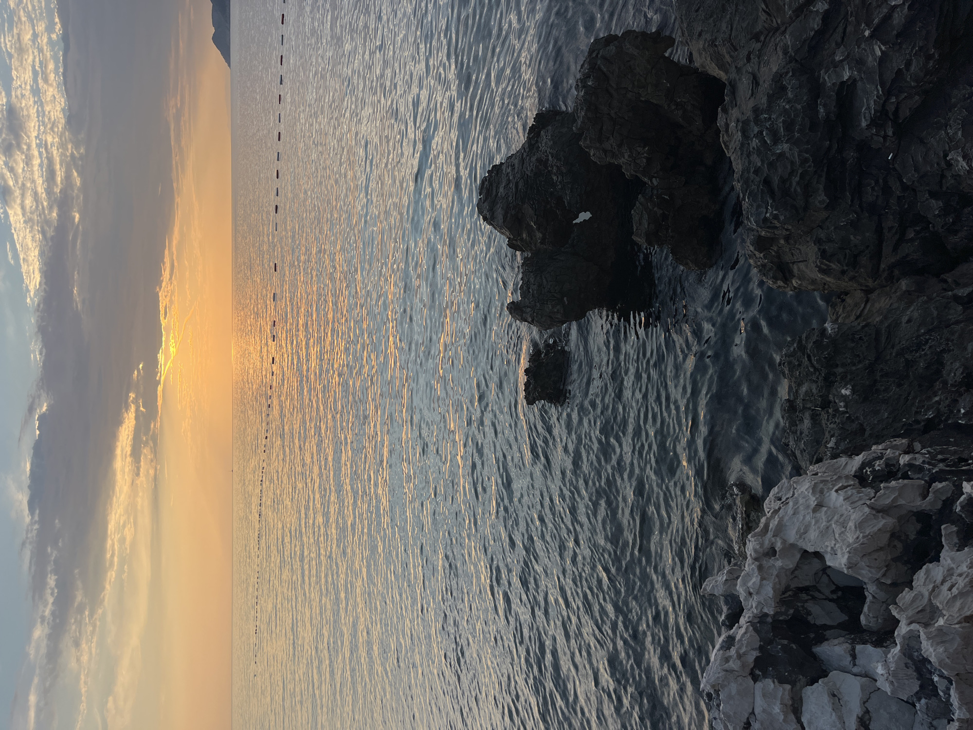 This is a vertical, eye-level shot of a calm sea meeting a rocky coastline at sunset. The sky on the left is a gradient of soft blues, greys, and oranges, with scattered clouds catching the last rays of sunlight. The sun itself is low on the horizon, casting a warm, golden reflection across the water, creating a shimmering pathway.
In the distance, a faint line of buoys or markers stretches across the water. The sea is a deep blue, with gentle ripples catching the light. On the right side of the image, dark, jagged rocks jut out of the water, their surfaces textured and wet. Towards the foreground, lighter, white rocks form the edge of the coastline, with rough, uneven surfaces and shadowed crevices.
The scene evokes a sense of tranquility and natural beauty, characteristic of a coastal sunset. The time of day is clearly late evening, as indicated by the sunset colors in the sky and the golden light on the water. The location appears to be a somewhat rugged and undeveloped coastline. There is no visible text in the image.