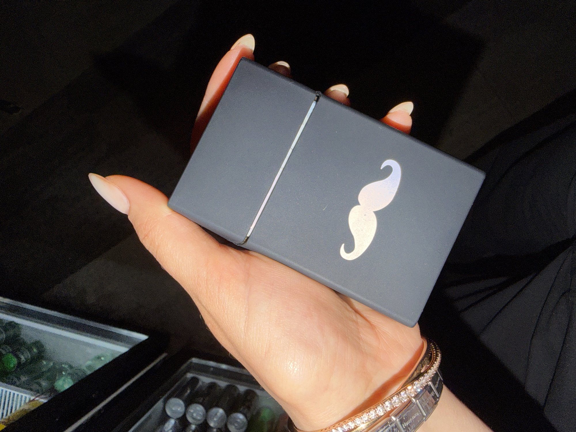 A close-up, high-angle shot captures a person's hand holding a dark gray cigarette case with a holographic mustache design on it. The hand, with long, almond-shaped light-pink nails, is adorned with a sparkling diamond bracelet. The background is dark and out of focus. To the left, a blurry glimpse of a cooler with drinks is visible. The image appears to be taken indoors in a dimly lit environment, potentially a bar or lounge. The exact location in the city is unknown.