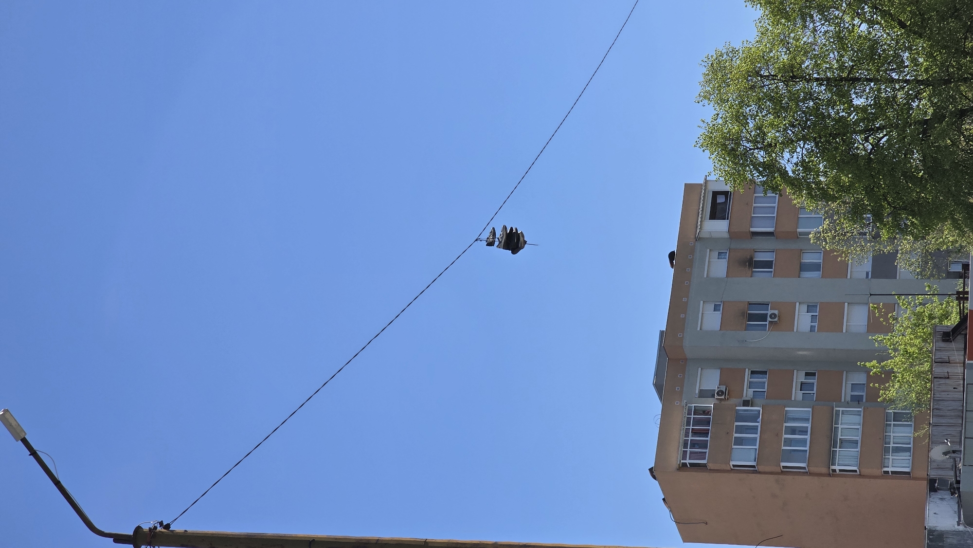 The image shows a pair of shoes dangling from a power line against a clear blue sky, with a multi-story building to the right and a street lamp to the lower left. The location is likely Blagoevgrad, Bulgaria. The shoes are dark-colored. The building is peach and gray with several windows, some of which have air conditioning units installed. Trees partially obscure the top of the building. The street lamp is gray and appears to be made of metal.