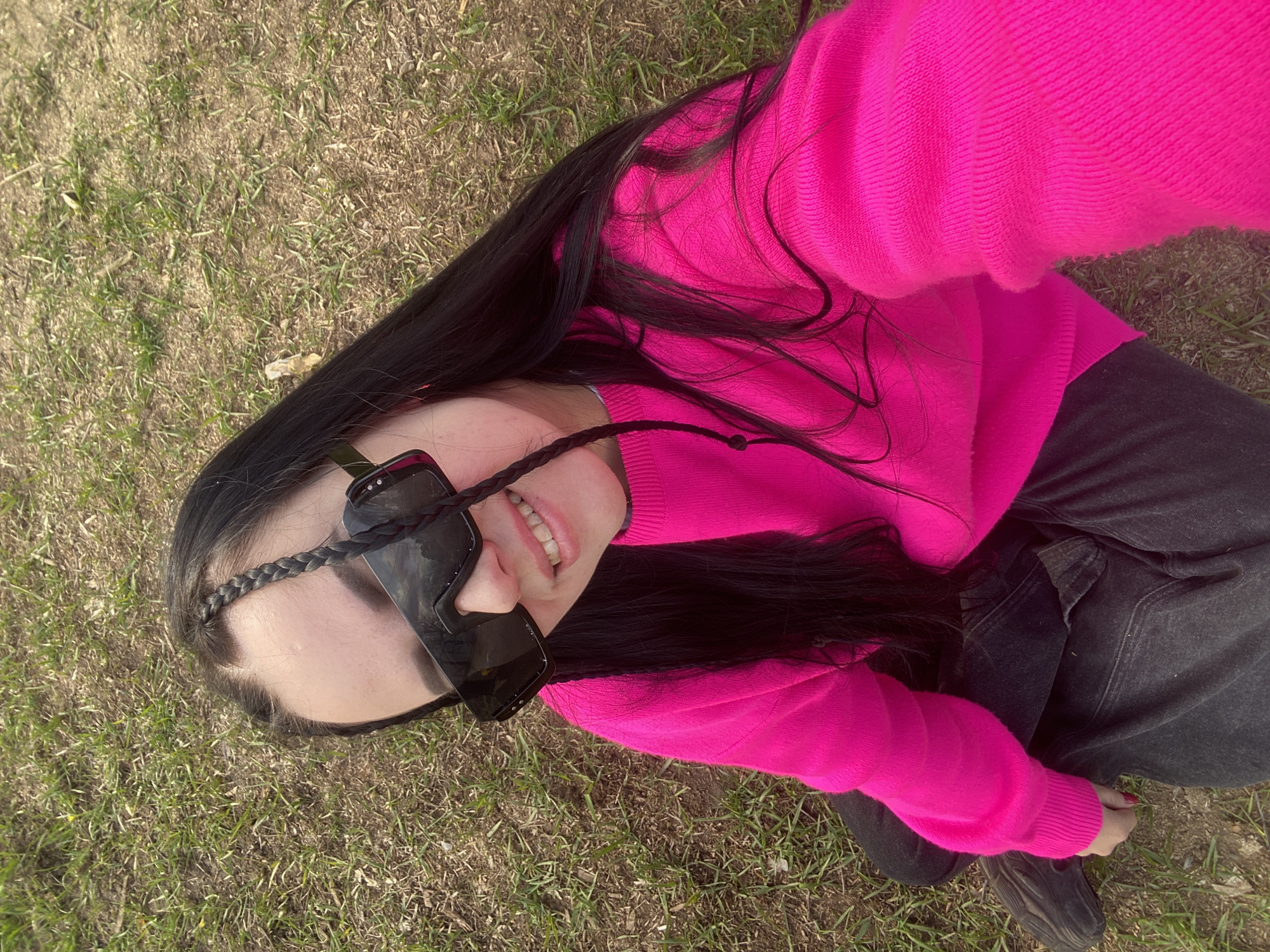 A young woman is lying on the grass in what appears to be a park. She has long, dark hair, with a braid running down the side of her face, and is wearing dark sunglasses. She's wearing a bright pink sweater and dark jeans. The ground is covered in grass, and she seems to be taking a selfie or having someone take a picture of her. The location is suggested to be Chaikovsky, Russia.