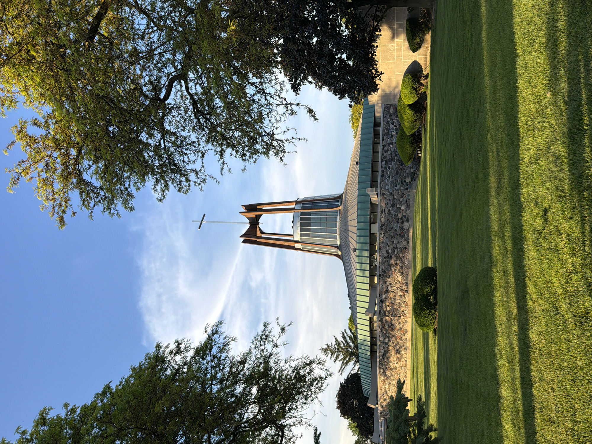 The image shows a low-angle view of a church, framed by trees, in Niles, United States of America. The church features a tall tower with a cross at its peak, set against a blue sky with wispy clouds. The building is constructed with a stone facade and a green roof. Manicured green lawns surround the building, with decorative bushes and trees nearby. Shadows cast by the trees streak across the grass.