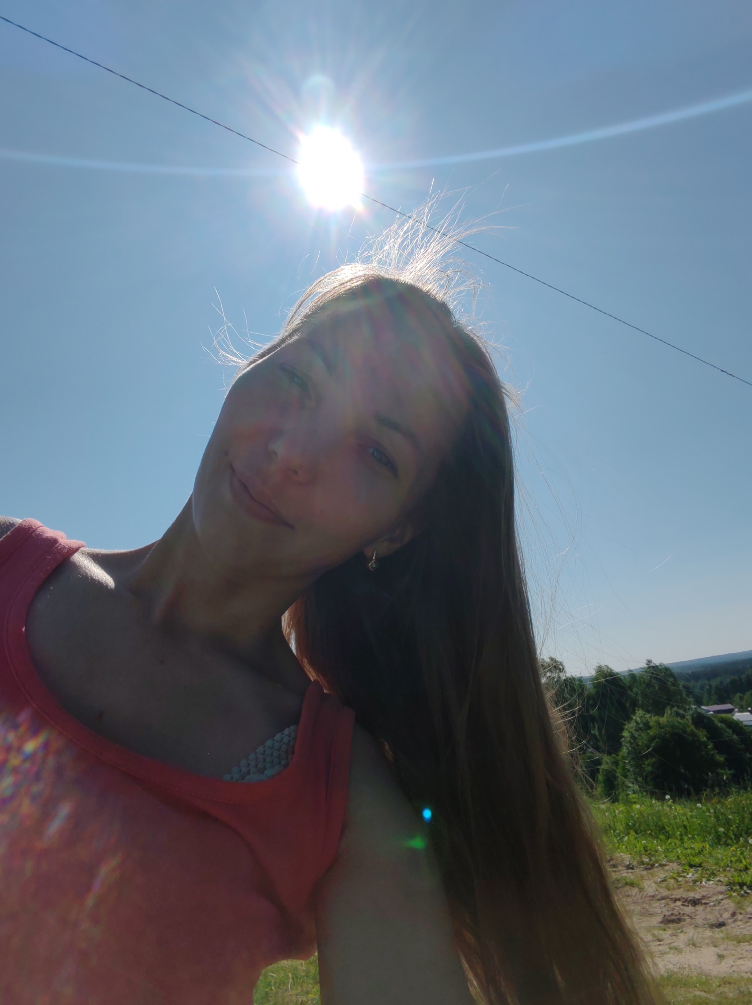 A woman is taking a selfie outdoors, with the sun shining brightly in the sky. She is wearing a pink tank top and has long, light brown hair. The background shows a green landscape with trees and some buildings in the distance, suggesting a rural or suburban setting possibly near Чухлэм, Russia. The lighting is strong, creating lens flare and highlights on her hair and skin.