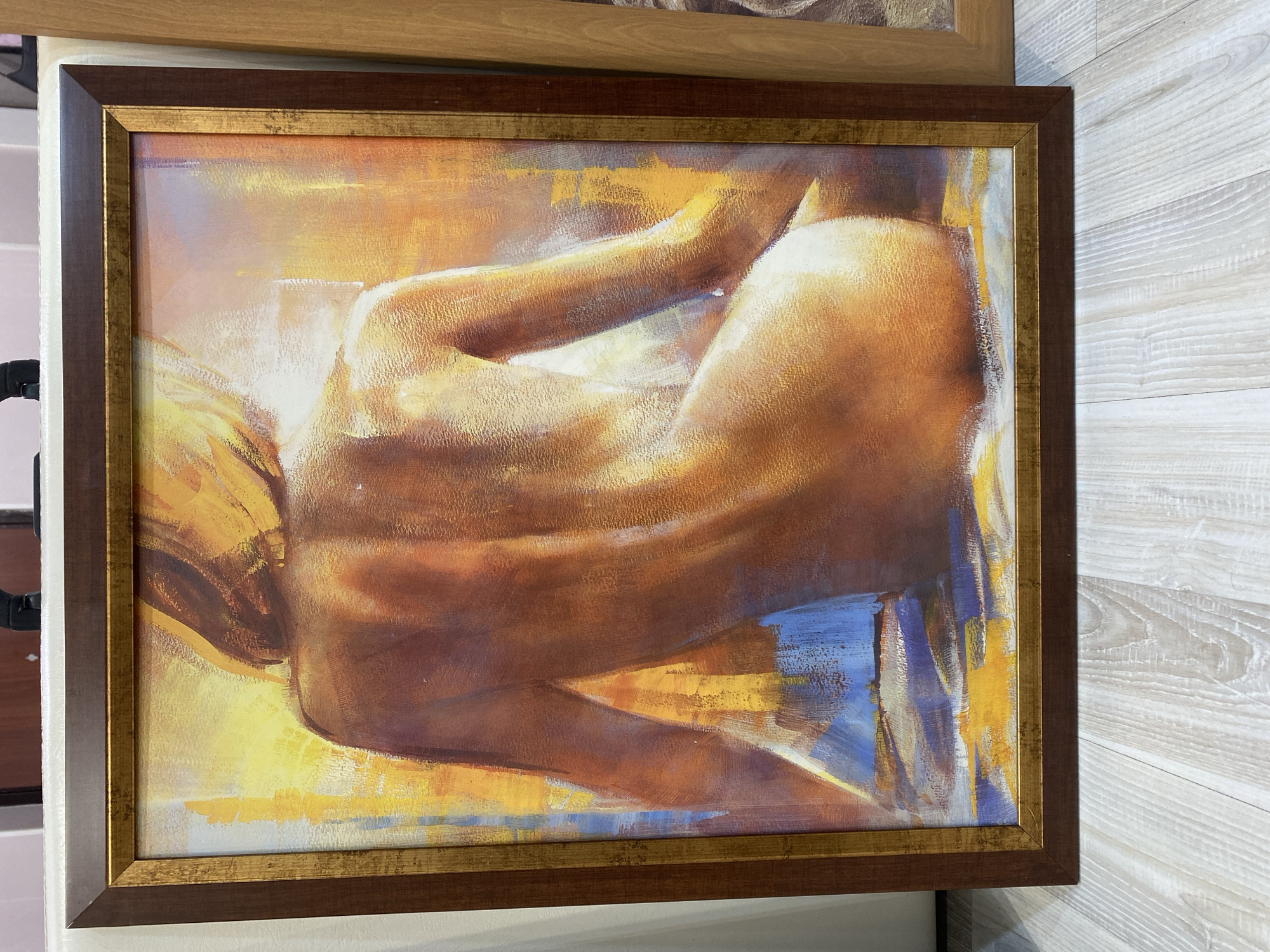 A framed painting of a nude figure is displayed in an interior setting. The painting is dominated by warm tones of yellow, orange, and brown, with some blue accents. The figure appears to be lying down or reclining, with their arm raised and the body curved. The painting is contained within a dark brown frame with a gold trim along the inner edge. The setting appears to be indoors with a light-colored wood-paneled wall partially visible behind the right side of the painting. The location and city are unknown.