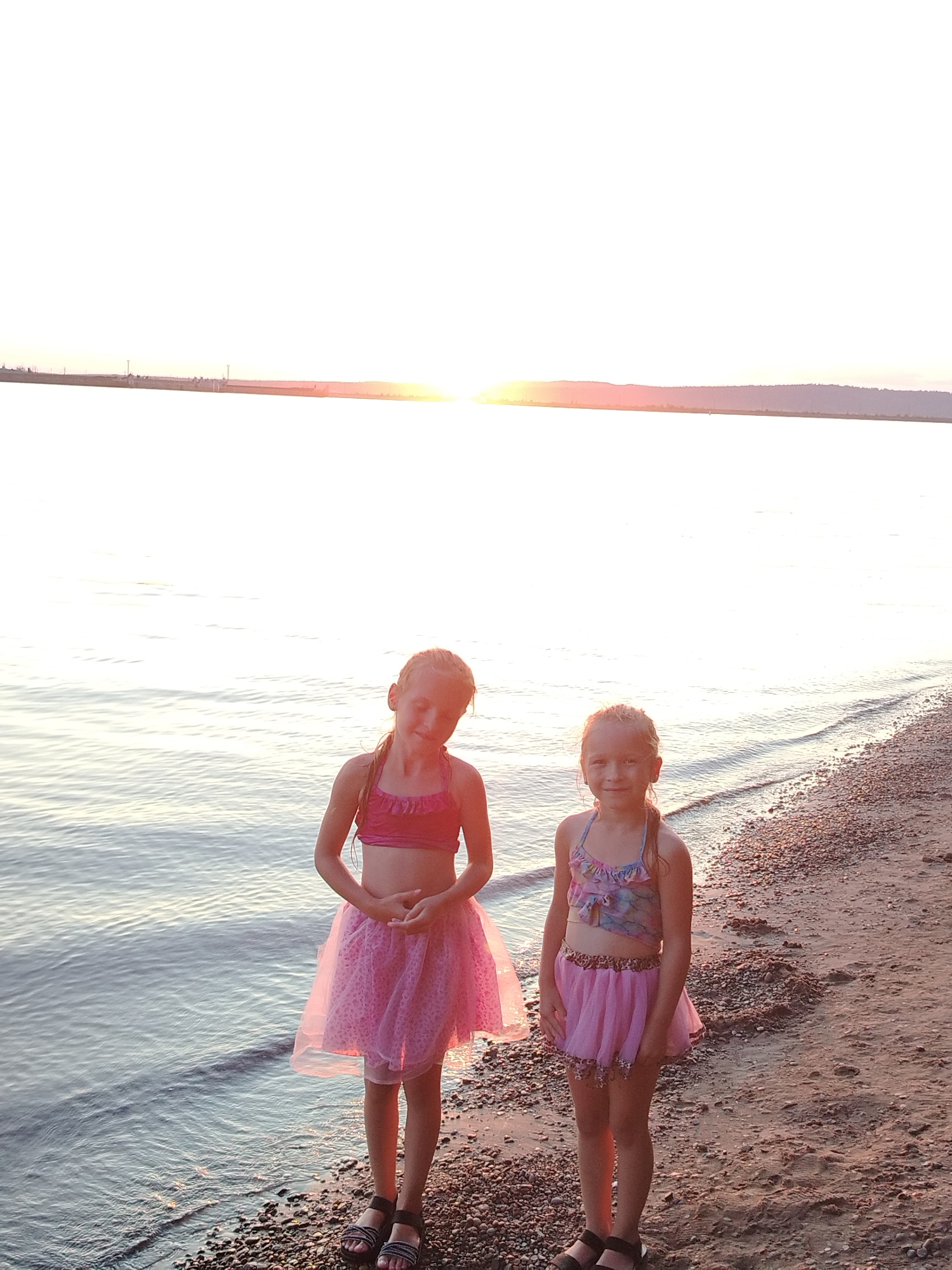 Two young girls are standing on a pebble beach next to a body of water during sunset. The girl on the left is wearing a pink two-piece swimsuit and a pink tulle skirt. The girl on the right is wearing a patterned swimsuit and a pink tulle skirt with a leopard print trim. Both girls are facing the camera. The sun is low on the horizon, casting a warm glow over the scene. The water appears calm with gentle ripples. In the background, the distant shore is visible with some indistinct structures and a hazy sky. The location is likely the Kama River embankment in Chaikovsky, Russia, given the typical sunset views over the water.