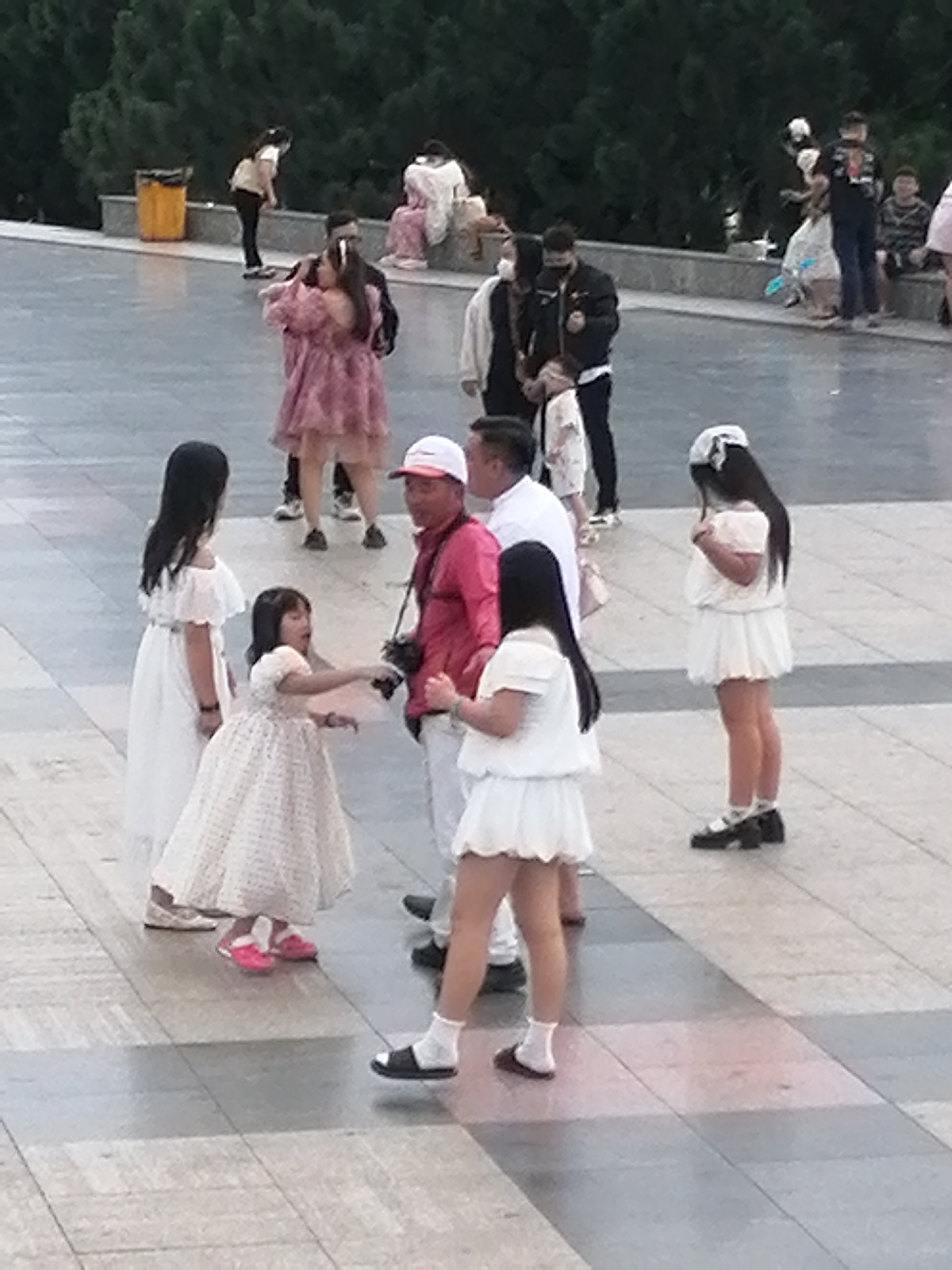 In Da Lat, Vietnam, on a paved public square surrounded by dense trees, a group of people are gathered. A man in a pink shirt and a white hat, holding a camera, stands in the center of the group. To his left, a young girl in a white dress reaches out, and a taller girl in a white dress stands behind her. To the man's right, a woman in a white dress with a white bow in her hair looks down. In the background, several other individuals are visible, some in casual attire and others wearing masks. The atmosphere appears to be relaxed, with people enjoying the outdoors.