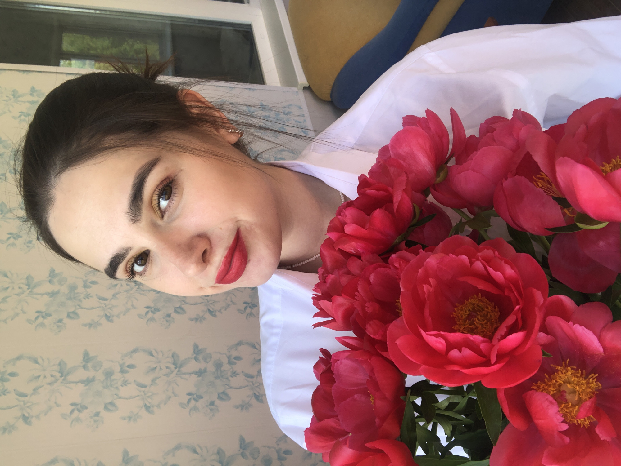 A young woman with dark hair pulled back in a bun and wearing a white collared shirt and a pearl necklace holds a bouquet of bright pink peonies. She is looking towards the camera with a slight smile, her lips painted red. Her right eyebrow is arched, and her eye makeup appears to include gold eyeshadow. She is wearing small, dangly earrings. The background consists of a floral-patterned wallpaper with light blue flowers on a pale background, and a window revealing a glimpse of yellow and blue furniture outside. The lighting suggests it is daytime. The image appears to be a selfie, taken indoors or in a covered outdoor space. There is no identifiable location in the city of Noviy, Russia, visible in the image.