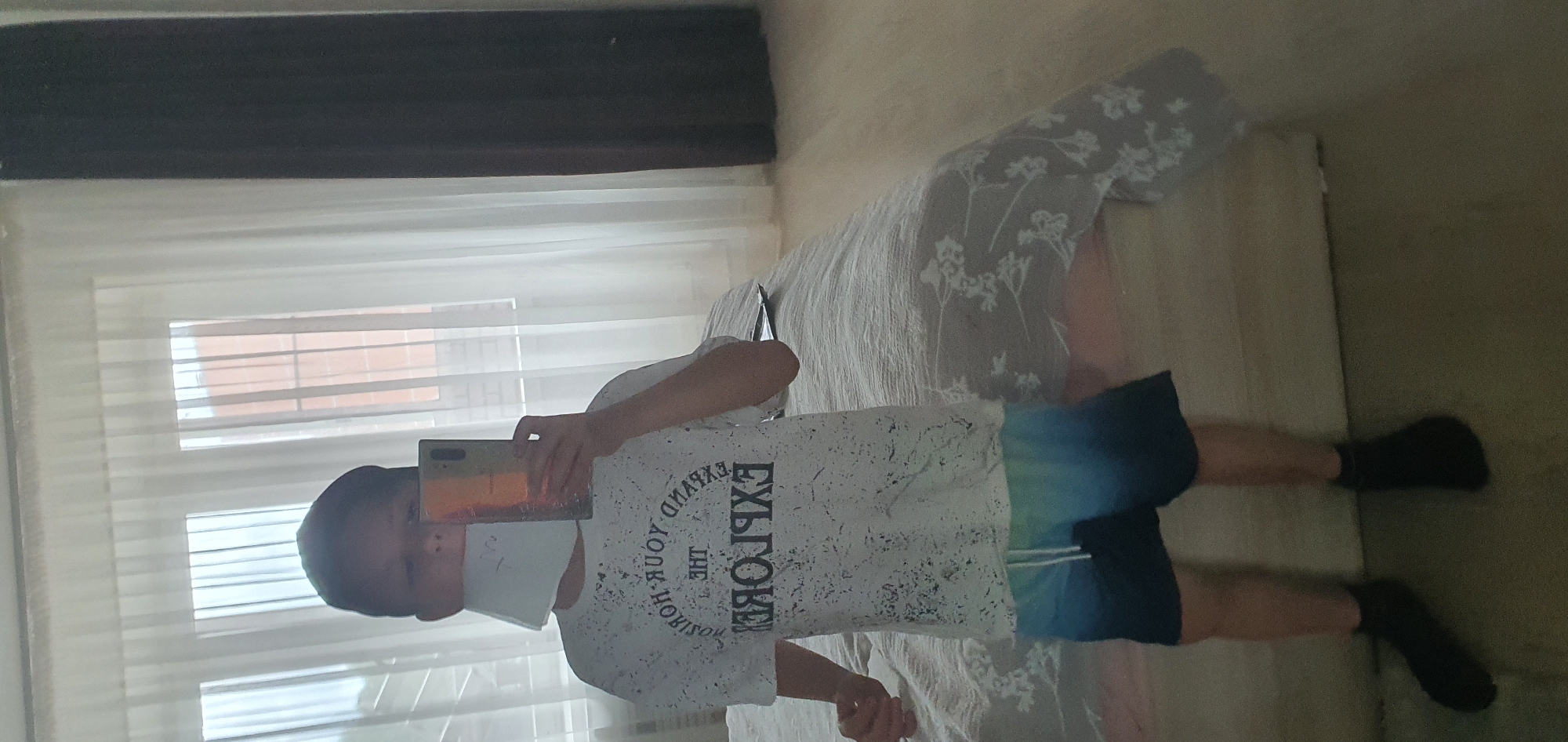 A person wearing a white t-shirt with the word "EXPLORE" and dark blue shorts is taking a selfie in a room. They are wearing a white face mask and holding a smartphone in front of their face, obscuring their features. The person is standing on a bed, which is covered with a floral-patterned blanket. Behind the person is a window covered with white curtains and blinds. The blinds are partially open, revealing a brick building outside. A dark brown curtain rod is visible above the window. The location is Chaikovsky, Russia.
