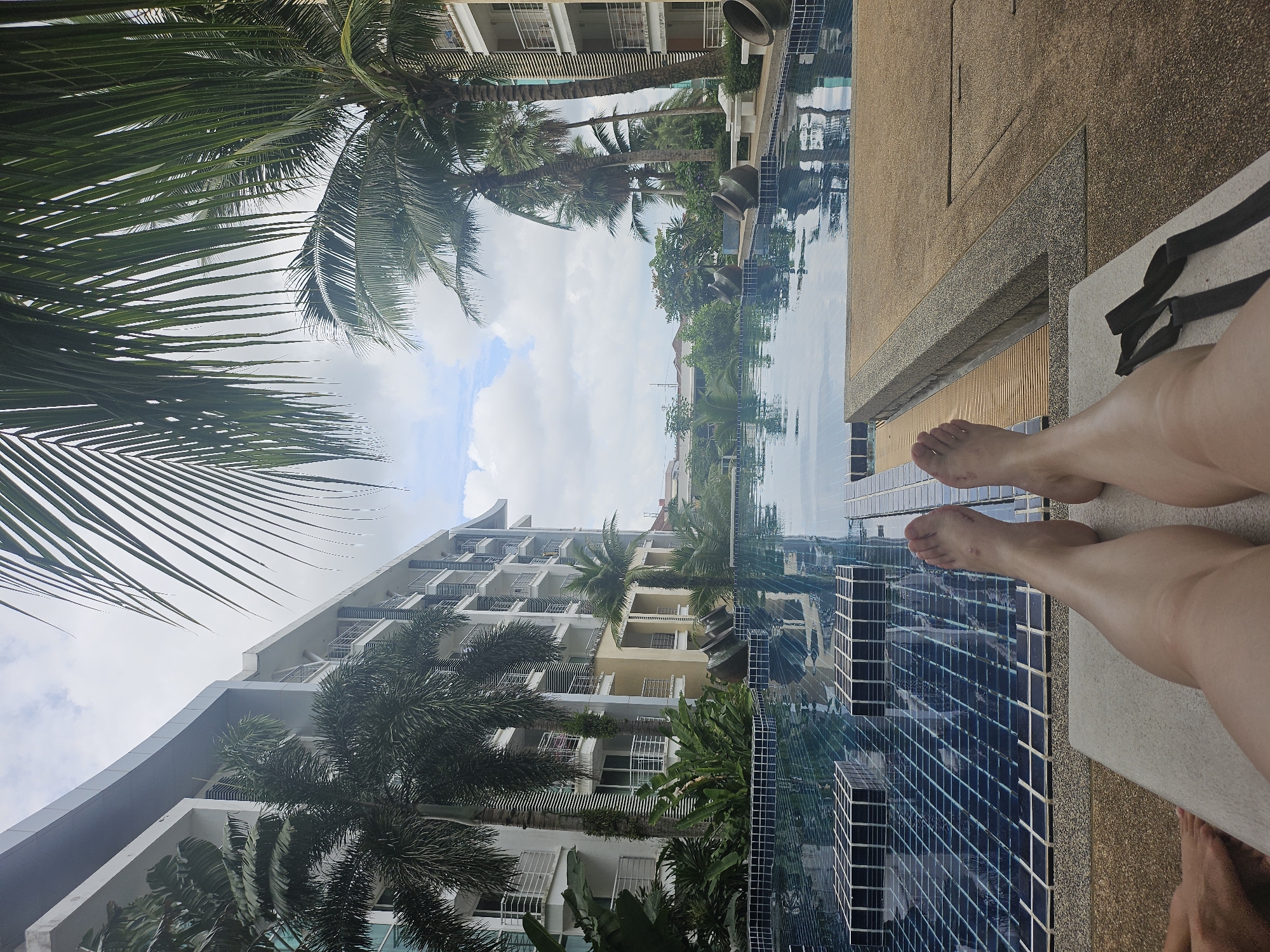 This image depicts a person's legs extended on a lounge chair, facing a swimming pool at an apartment complex in Kathu, Thailand. The pool features blue tiled flooring and is surrounded by a stone deck. Several palm trees are visible, along with a multi-story apartment building. The sky is overcast with scattered clouds.