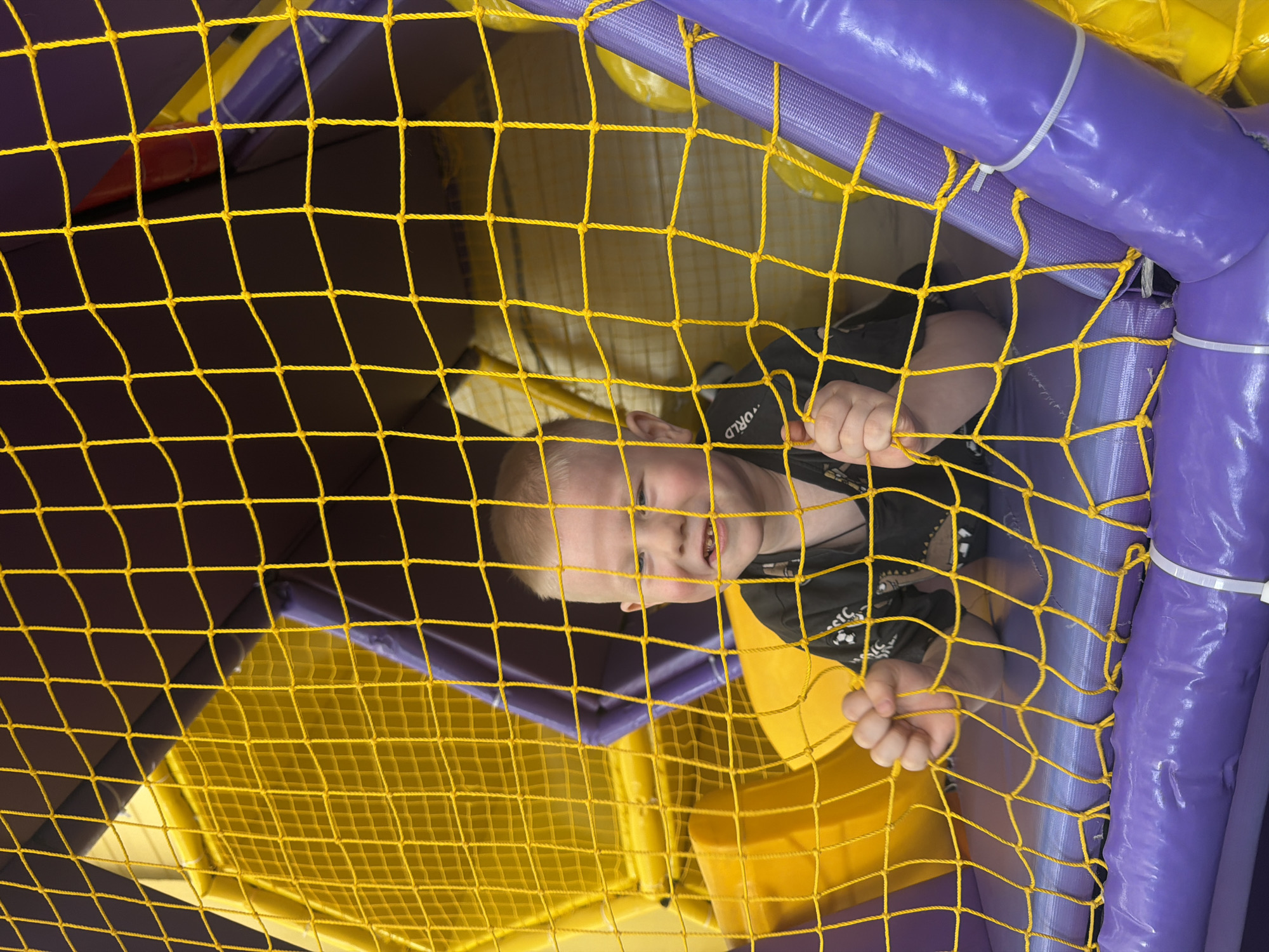 A child is seen through a yellow rope net structure within a purple and yellow padded play area. The child is wearing a dark colored t-shirt with a light colored design on the front. The play area appears to be indoors, likely in a city such as Chaikovsky, Russia. The child's hands are visible gripping the yellow net.