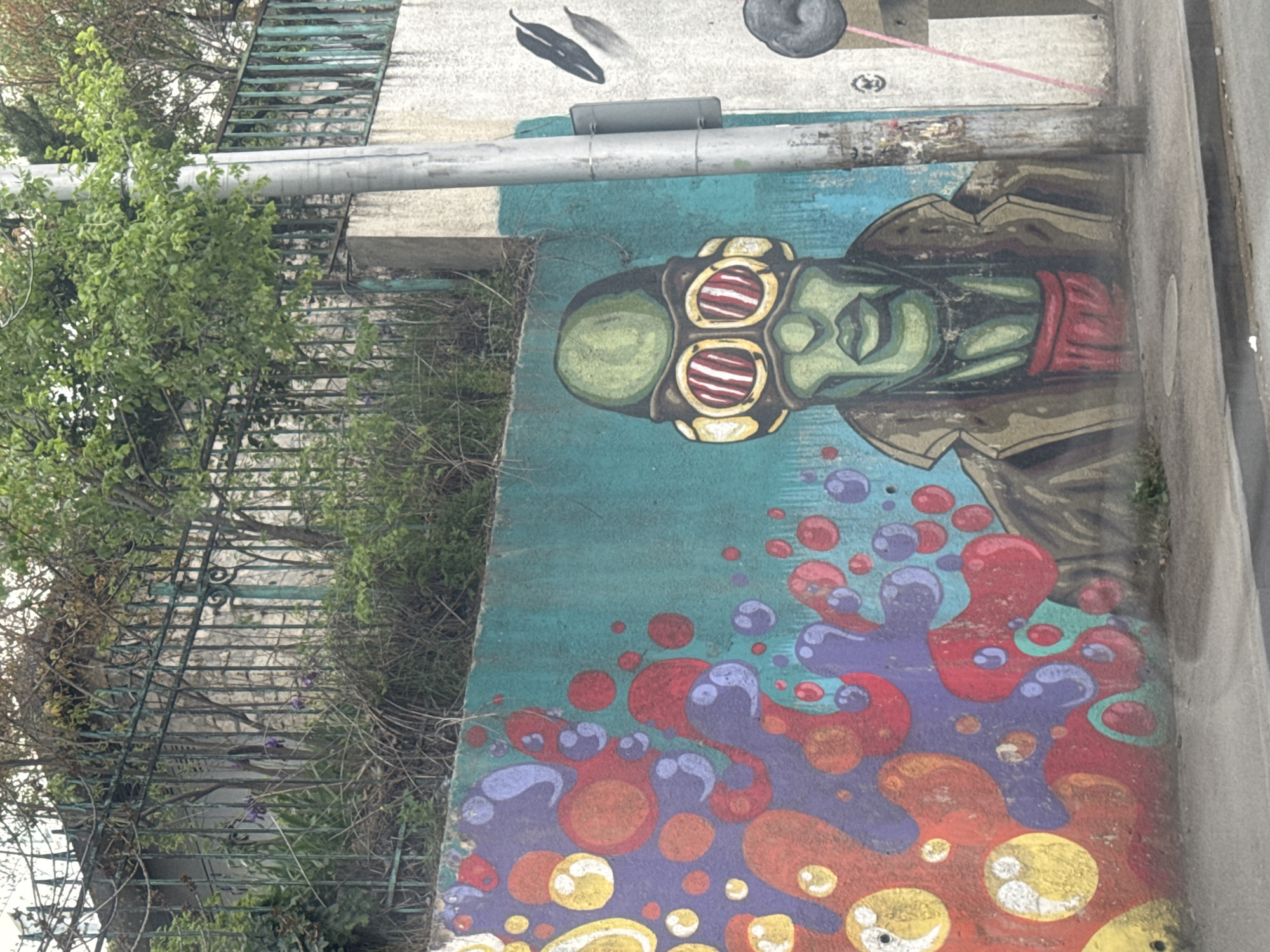 A vibrant street art mural is depicted on a wall in Tbilisi, Georgia, viewed from a slightly elevated perspective. The main subject of the mural is a stylized human-like figure with green skin, seen from the chest up. The figure wears large, multi-lens goggles with red and yellow horizontal stripes, and a brown or olive-green jacket with a red inner collar or scarf. Its expression appears neutral or contemplative with closed lips.
Below the figure, the mural transforms into a dynamic abstract design featuring numerous colorful, organic shapes resembling bubbles or liquid splatters. These elements are rendered in shades of red, purple, orange, and yellow, set against a teal background that transitions to a darker blue-green towards the top.
To the left of the mural, a dark metal fence, heavily overgrown with various green foliage and dry vines, extends downwards. A long, light grey metal pipe is mounted horizontally across the concrete structure above the mural. The concrete wall above the pipe is light-colored, showing some minor wear and faint, unidentifiable markings. No discernible text is visible on the mural or its surroundings. The scene is illuminated by natural daylight, suggesting it is daytime, with an overall soft light rather than direct sunlight. The image captures an intersection of urban infrastructure, nature, and street art.