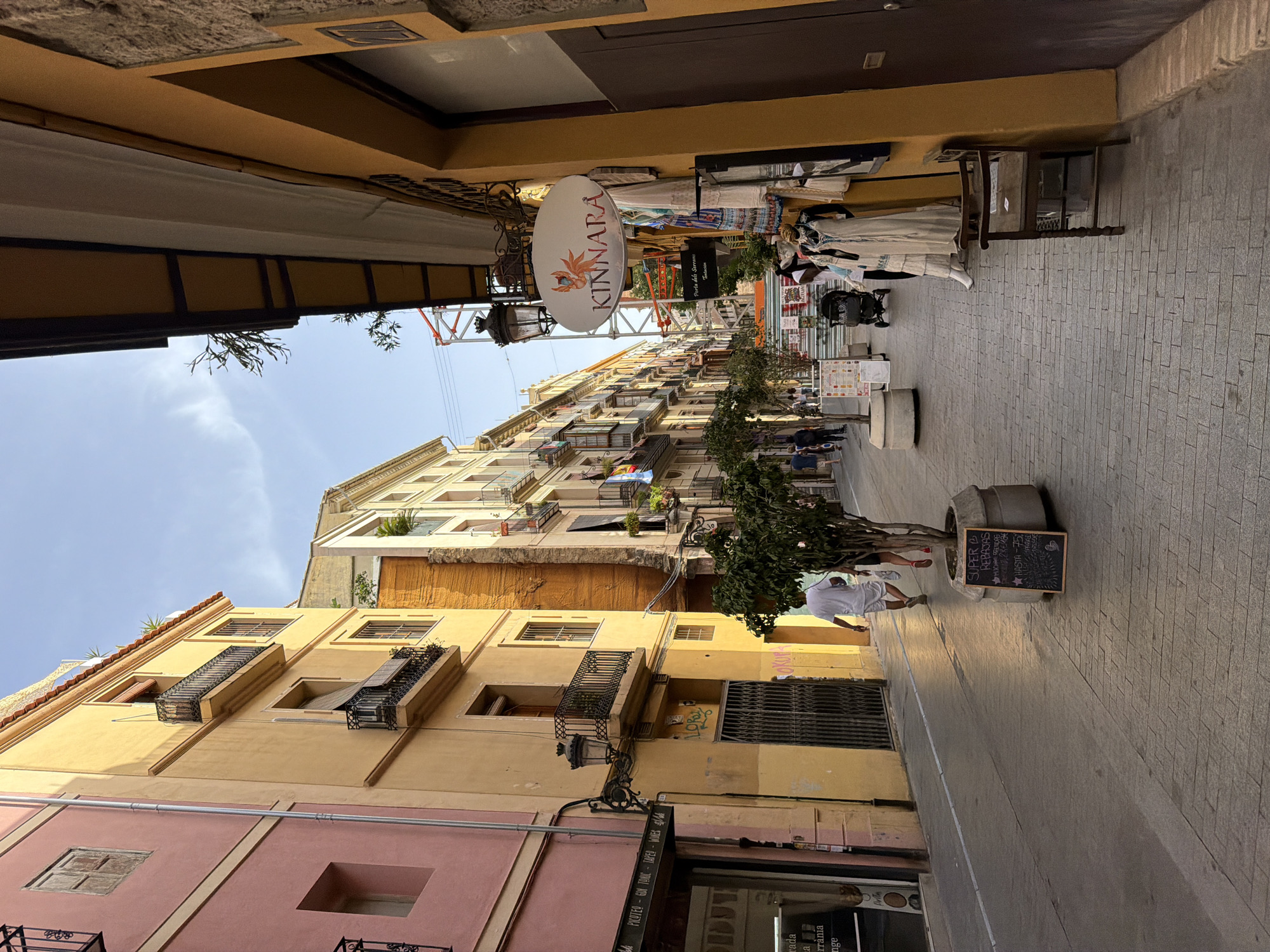 The image shows a narrow, pedestrianized street in a European city, likely in Southern Europe given the architecture and warm color palette. The street is lined with multi-story buildings painted in shades of pink and yellow, with balconies and shuttered windows. The pavement is made of grey cobblestones. On the right side of the street, a yellow building has a store with clothing displayed outside, including mannequins wearing dresses. A sign for "KINNARA" is visible above the store. Further down the street, there are more buildings, some with laundry hanging from balconies, and people walking. A man in a white shirt and shorts is walking away from the camera on the right side of the street, next to a potted tree. A small blackboard sign in front of the tree displays text and stars. On the left side, a pink building has a store with a sign that reads "PICOTEO - GIN TONIC - TAPAS - WINES". The sky is blue with some wispy clouds. A construction crane is visible in the background. The scene appears to be during daylight, with the sun casting shadows on the street.