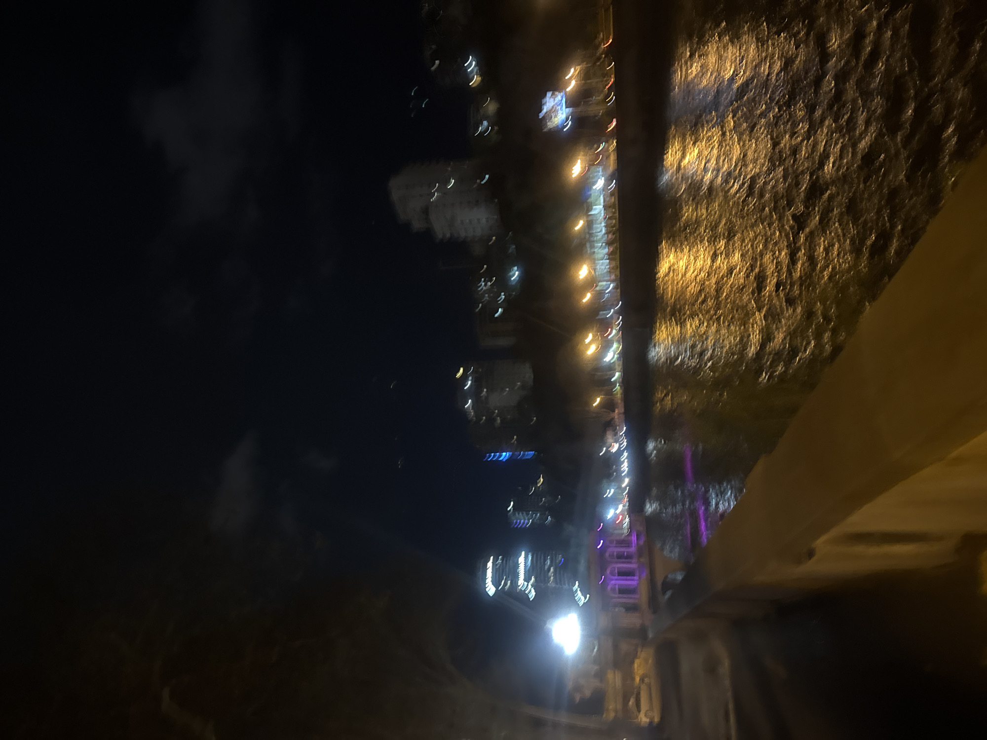 The image depicts a night scene in Tbilisi, Georgia, likely taken from a bridge. The right side of the image shows a concrete structure, possibly the bridge's barrier, with textured, golden light reflecting on its surface. A row of lights stretches along the bottom half of the picture from left to right and can be spotted on buildings on the left side in the far distance. There is a dark body of water in the middle. The background features a dark sky with clouds.