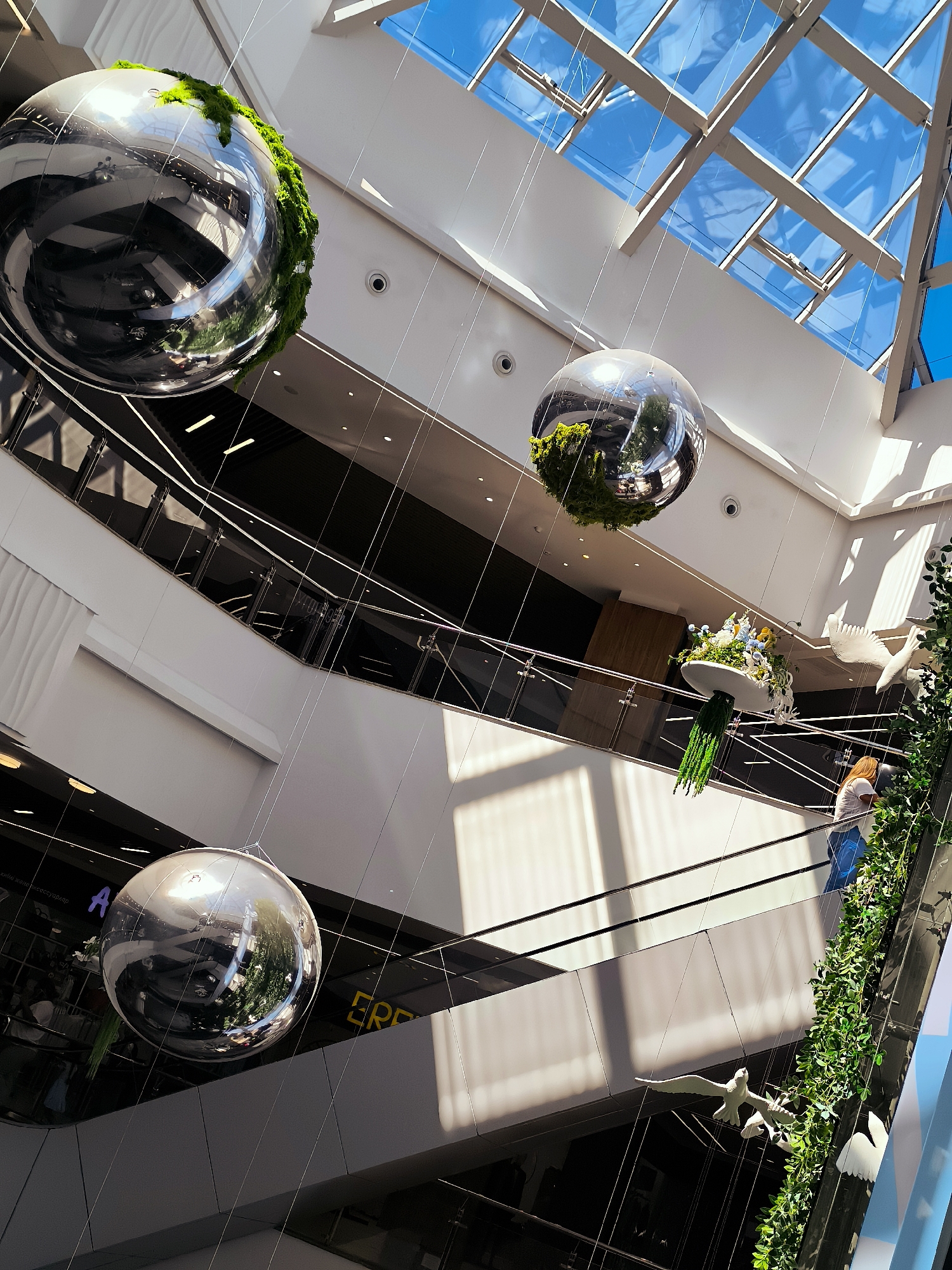 This image captures the interior of a multi-level modern building, possibly a shopping mall or atrium, with a distinctive glass ceiling allowing natural light to stream in. The scene is dominated by large, reflective metallic spheres suspended from above, some adorned with mossy green accents.
Below the spheres, multiple levels of walkways and escalators are visible, with glass railings and minimalist architectural designs. Several decorative elements are also suspended: a floral arrangement in a white dish with green fringe, and white bird-like sculptures. A person is seen on an escalator, wearing a blue shirt and white pants, looking towards the greenery. Another person is partially visible on a higher level, wearing a white shirt and blue jeans, interacting with the hanging greenery.
The lighting suggests it is daytime, with bright sunlight filtering through the glass ceiling, creating strong shadows and highlights that accentuate the modern architecture. The overall mood is one of contemporary design and a bright, airy atmosphere.
Visible text includes what appears to be a purple "A" on a dark surface on a lower level, and a yellow "ERP" on a dark surface on another lower level. The overall impression is an aesthetically pleasing and spacious indoor environment with unique artistic installations.