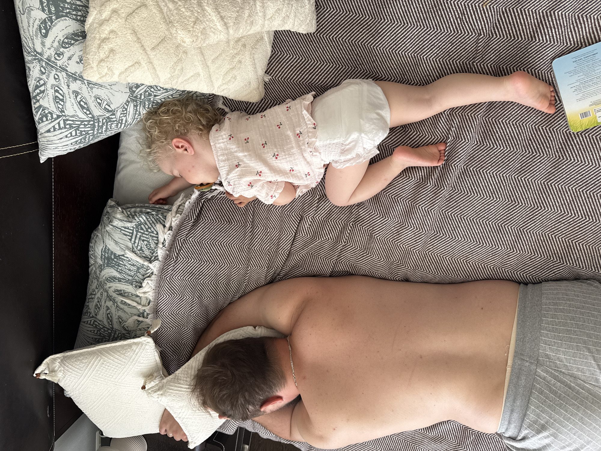 An overhead, high-angle shot shows two people sleeping on a bed. The person on the left is an adult male lying on his stomach, facing away from the camera. He is wearing grey striped pajama bottoms and a silver chain necklace. The person on the right is a young child, likely a toddler, lying on their side, facing away from the camera. The child has blonde, curly hair and is wearing a pink t-shirt with a cherry print and a diaper. The child has a pacifier in their mouth. Both are sleeping on a bed with a brown and white chevron patterned blanket. There are several pillows on the bed, including a white textured one, a light blue and white botanical print pillow, and another white textured pillow. The scene is indoors, likely a bedroom. No specific location in Vladimir, Russia is indicated.