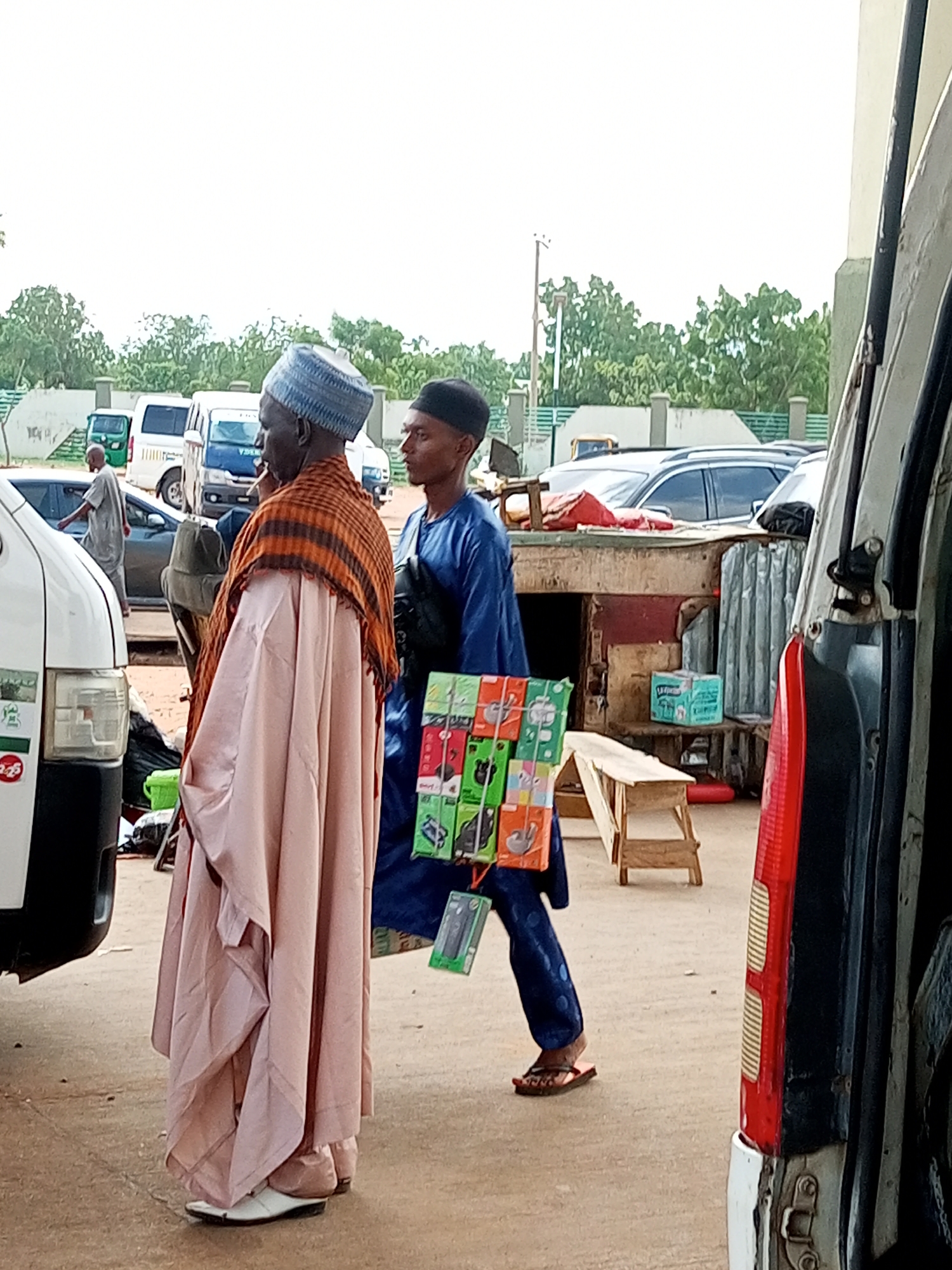 This image depicts an outdoor scene in the city of Gombe, Nigeria, likely a marketplace or a roadside vending area. Two men are the primary subjects. The man on the left is older, wearing a light pink robe and an orange and brown checkered scarf draped over his shoulders. He has a blue and white patterned cap on his head and white shoes. The man on the right is younger, dressed in a blue robe and a black cap. He is holding a rack of items, which appear to be phone accessories like chargers and cases, displayed on colorful packaging.
In the background, several vehicles are visible, including white vans and a small three-wheeled vehicle. Some of these appear to be parked or possibly for sale or hire. There is a wooden stall or kiosk in the middle ground with some goods displayed, including a blue box of what looks like diapers. The ground is paved, and the overall environment suggests a bustling commercial area. The sky is overcast.