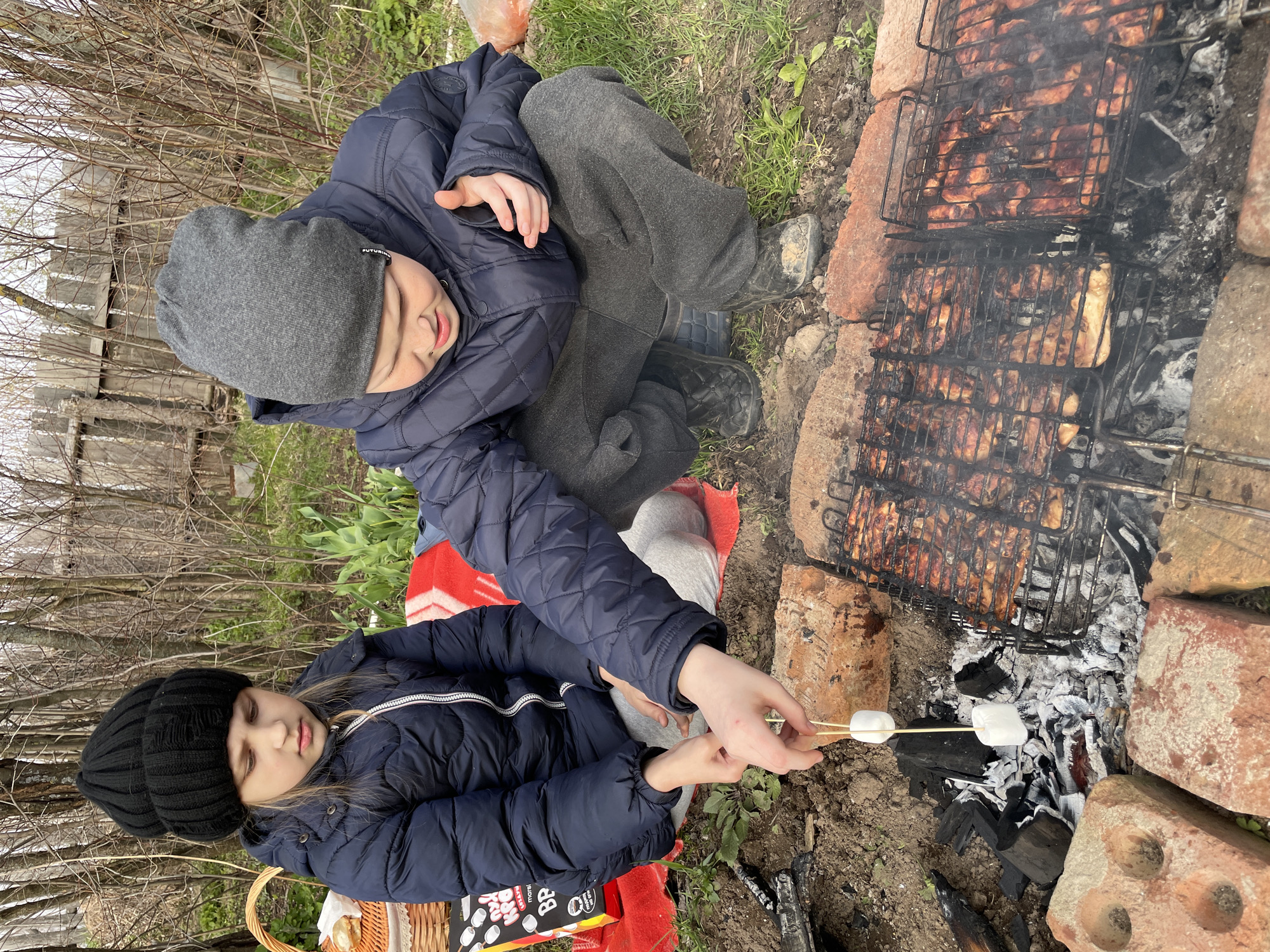 Two children, one wearing a black beanie and the other a grey beanie, are sitting near a brick barbecue grill. The girl in the black beanie is roasting marshmallows over the embers of the grill, while the boy in the grey beanie sits beside her. The grill contains cooked meat. The location appears to be outdoors in a natural setting, consistent with Балдейка, Russia. A red blanket is visible.