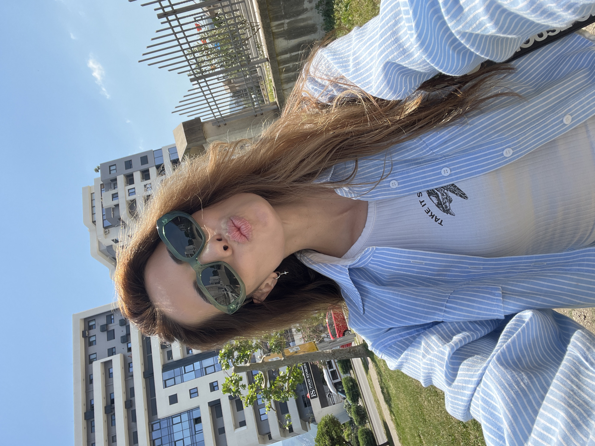 A woman with light brown hair and wearing green-framed sunglasses is posing for a selfie. She is puckering her lips towards the camera. She wears a white ribbed top underneath a blue and white striped shirt. A thin, hoop-style earring is visible in her ear. Buildings with multiple floors are visible in the background, as well as green grass and a gray sidewalk. The scene is located outdoors in an urban area in the city of Bar, Montenegro.