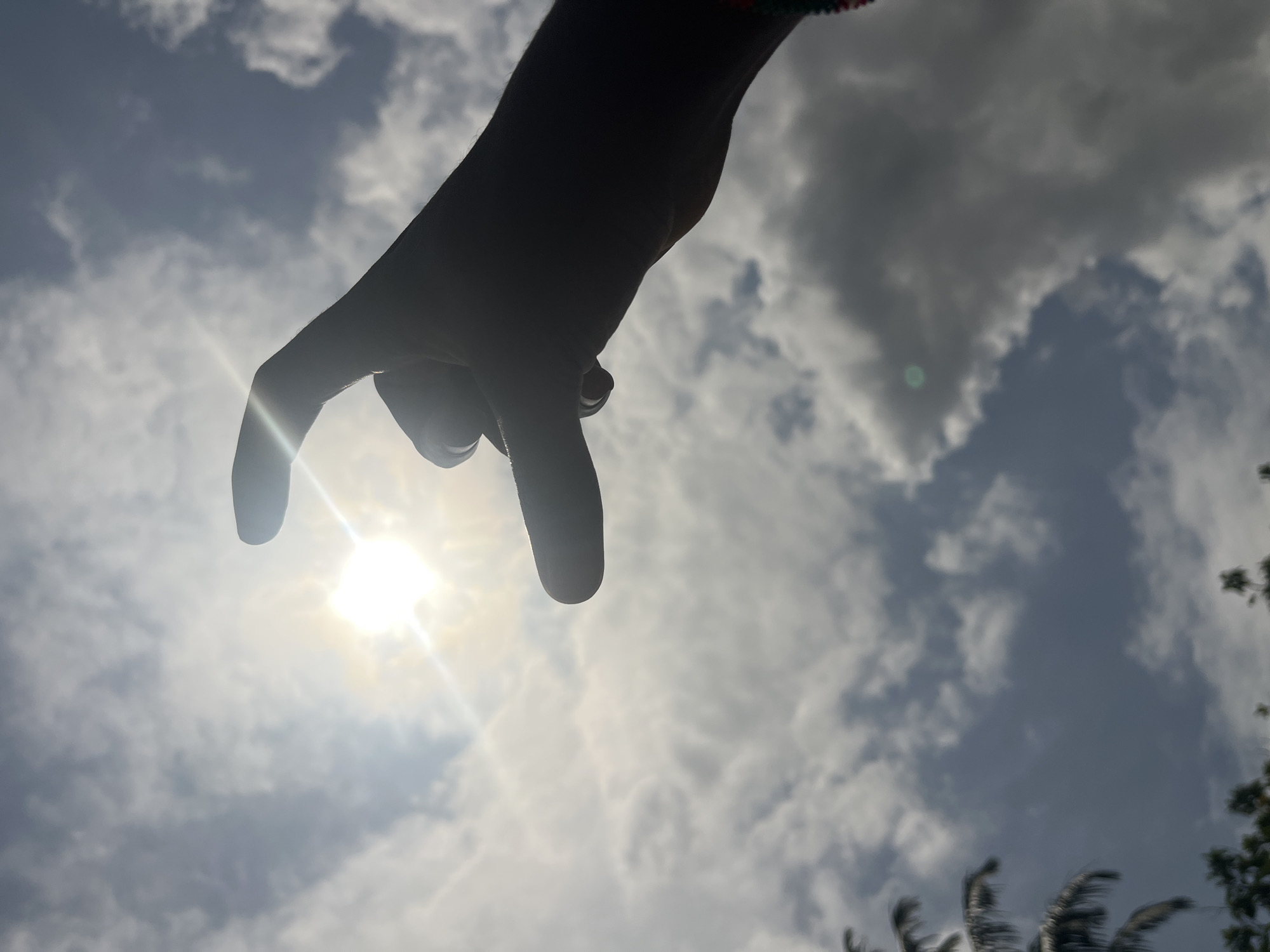 The image presents a dramatic low-angle shot of a person's hand silhouetted against a bright, cloudy sky. The hand, belonging to an adult, is positioned in the upper central area, held up or reaching down, with fingers slightly bent. A faint, colorful bracelet with red and green elements is visible on the wrist at the top edge of the hand.
The sun is directly behind and below the hand, creating an intense lens flare that obscures details but highlights the hand's dark outline. The sky is a mix of bright white and grey cumulus clouds, with hints of blue sky showing through. In the bottom right corner, blurred, dark green foliage, possibly palm fronds, is visible, suggesting a natural outdoor setting.
The image conveys a sense of brightness and strong contrast. The time of day is clearly daylight, likely midday or early afternoon due to the high sun position and intensity. The weather is sunny with significant cloud cover. The presence of palm-like foliage aligns with the given location context of Ikot Ekpene, Nigeria, which has a tropical climate. There is no visible text in the image.