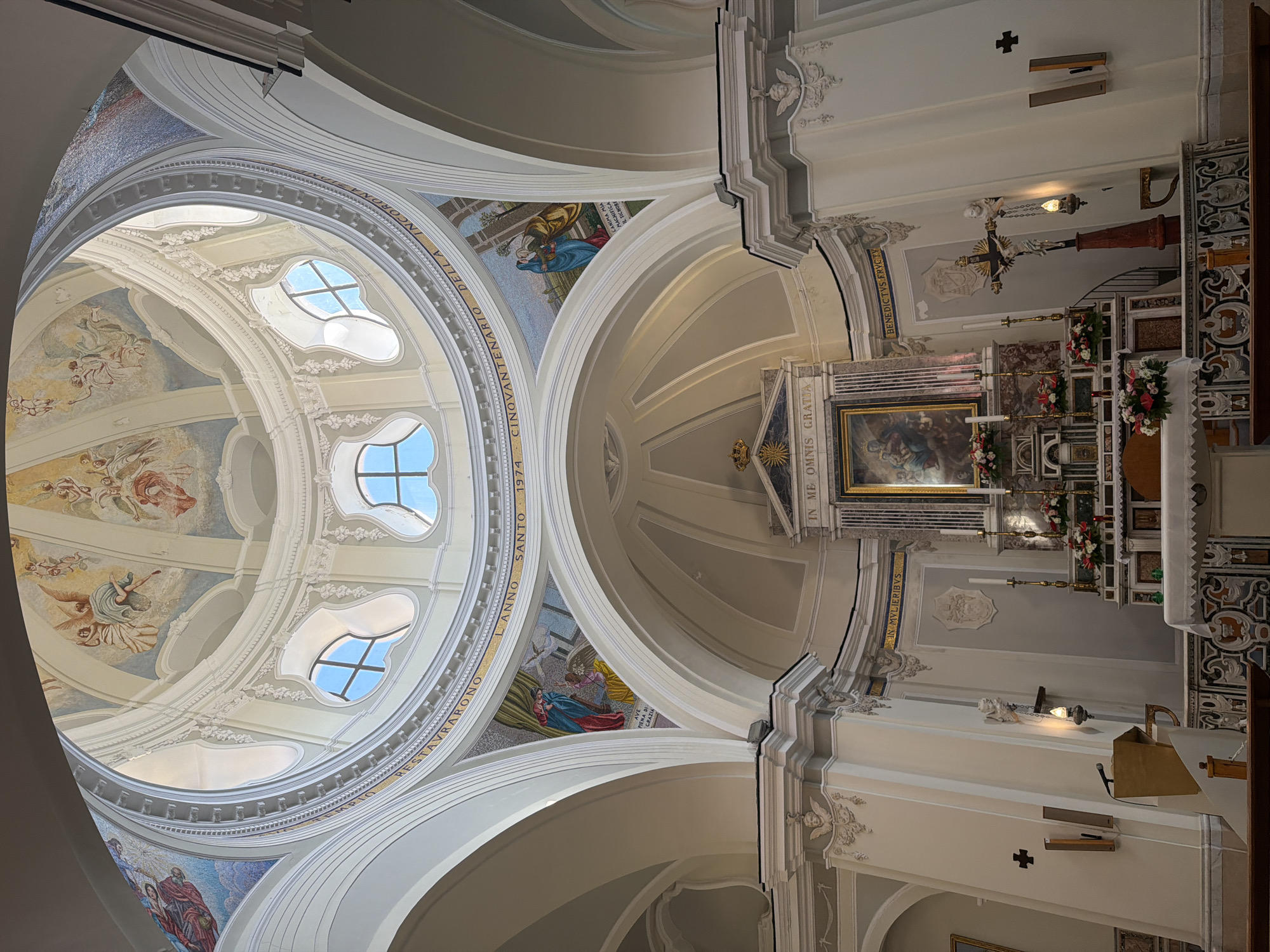 This is an interior shot of a church in Procida, Italy. The camera is pointed upwards, revealing two domes with murals of religious figures and symbols. The largest dome has multiple windows with a unique quatrefoil shape. Mosaics are displayed at the base of the domes.
Below the domes is a Catholic altar, covered with a white cloth, flowers, candles, and various religious artifacts. A large, ornate painting in a gilded frame hangs above the altar. A small cross is mounted on the wall above the altar. Various architectural details adorn the walls, including carvings and decorative moldings. A cross is mounted on the wall to the right of the altar.
The inscriptions on the domes and altar are in Latin, which references the church's history and religious significance.