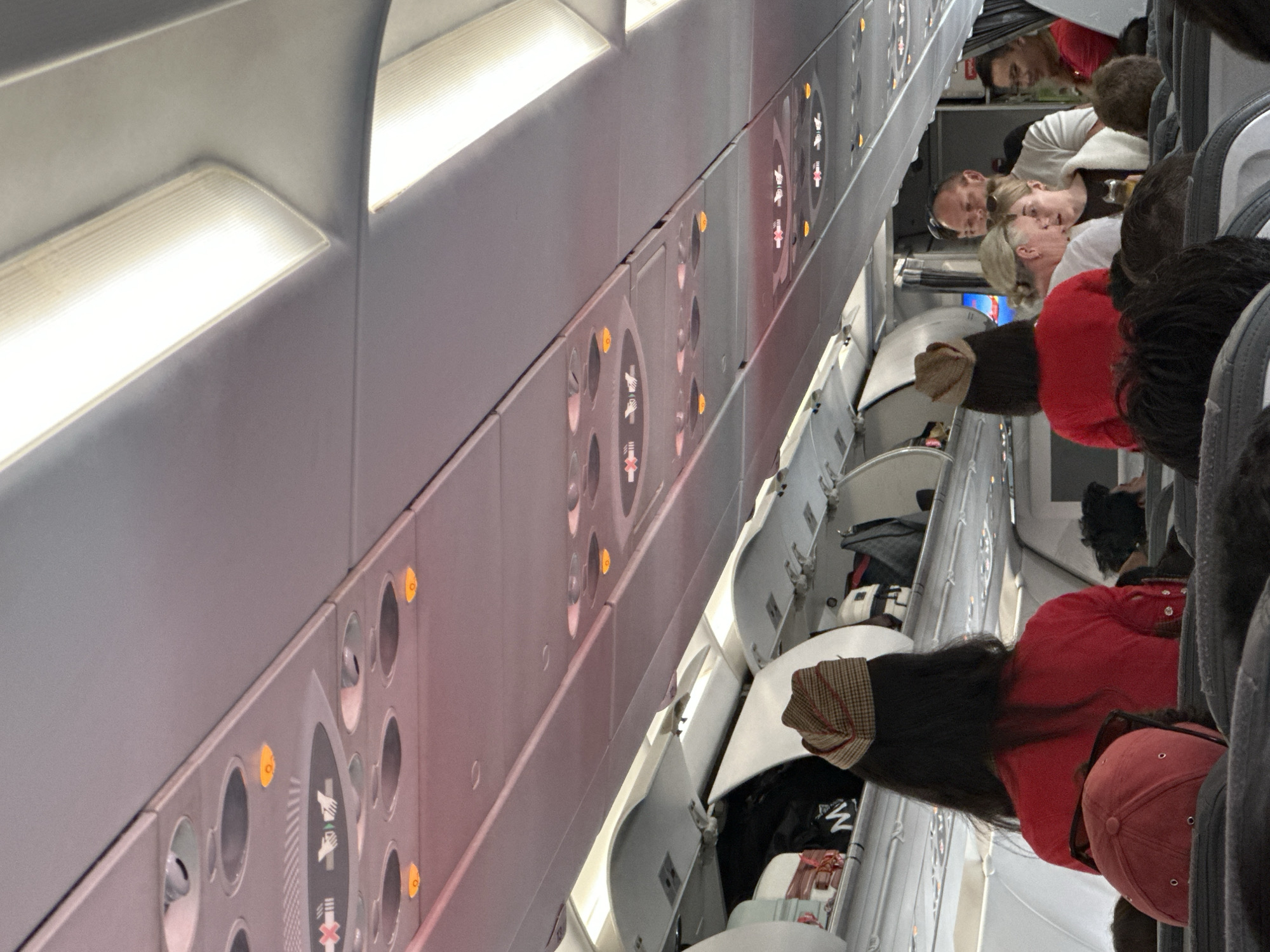 This image depicts the interior of an airplane cabin. The perspective is from a passenger's seat, looking forward and upwards towards the ceiling panels and overhead bins. The ceiling features rows of illuminated circular lights and wider rectangular light fixtures. Signs with icons, such as a crossed-out hand and two hands reaching, are visible on the panels. The overhead bins are open in several locations, revealing luggage and bags.
In the foreground, passengers are seated, with the back of a person wearing a red shirt and a patterned hat visible in the left half of the frame. Further down the aisle, several people are standing and interacting, including a woman with blonde hair and another person wearing glasses. A flight attendant, identified by a red uniform and a patterned hat, is also visible.
The scene suggests a typical commercial flight environment. While the image is not explicitly located in Kuta, Indonesia, the context of an airplane cabin implies travel, and Kuta is a well-known tourist destination in Indonesia, making it a plausible origin or destination.