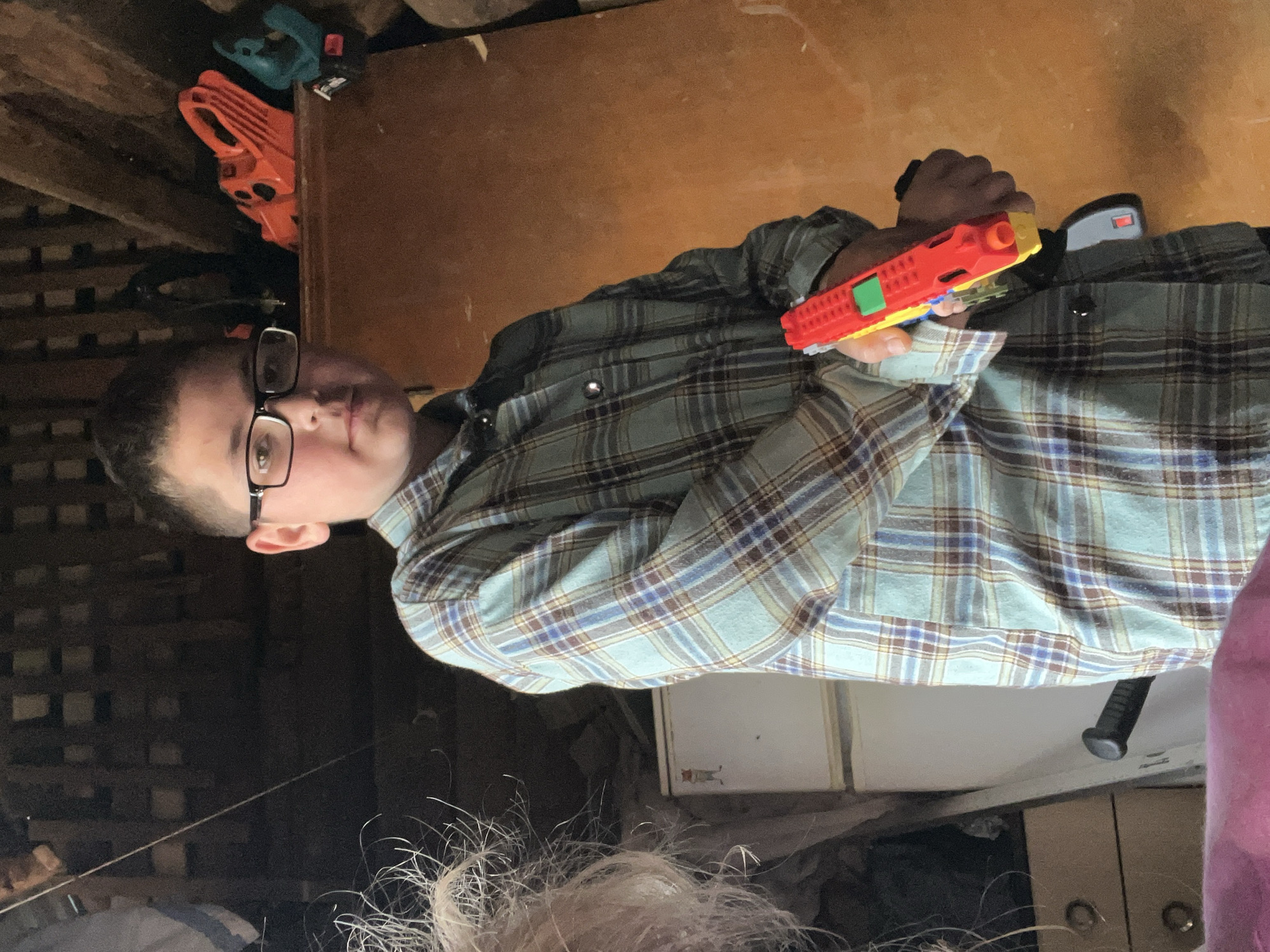 A young male wearing glasses and a plaid shirt stands in a dimly lit room, holding a red and yellow toy gun. He is positioned in the center of the frame, with his body turned slightly to his left. The background is cluttered with various items, including what appears to be storage shelves filled with tools and objects, a white appliance, and wooden walls. The overall setting suggests an indoor, possibly workshop or storage area, environment. The location in the city of Фоки, Russia cannot be determined from the visual information provided in the image.