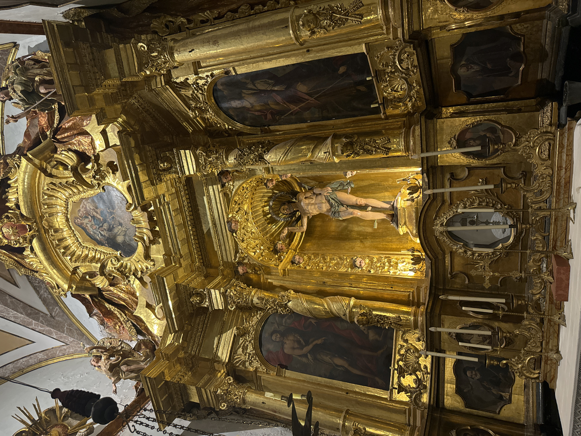 The image shows an ornate, gilded altarpiece within a church in Sóller, Spain. The altarpiece is heavily decorated with sculptures, paintings, and architectural elements. In the central upper section, there is a painting depicting the Virgin Mary and Child, framed by gilded carvings. Below this, a large sculpture of Jesus Christ on the cross is the focal point, with his body illuminated. Flanking the central crucifix are two vertical paintings. The left painting depicts a male figure in a red robe, possibly Saint John the Baptist. The right painting shows a figure in armor holding a sword. The lower section of the altarpiece features several smaller oval portraits of bearded male figures, and ornate candle holders with lit candles. The entire structure is made of gilded wood, with intricate carvings and decorative details throughout, suggesting a significant religious or historical site in Sóller.