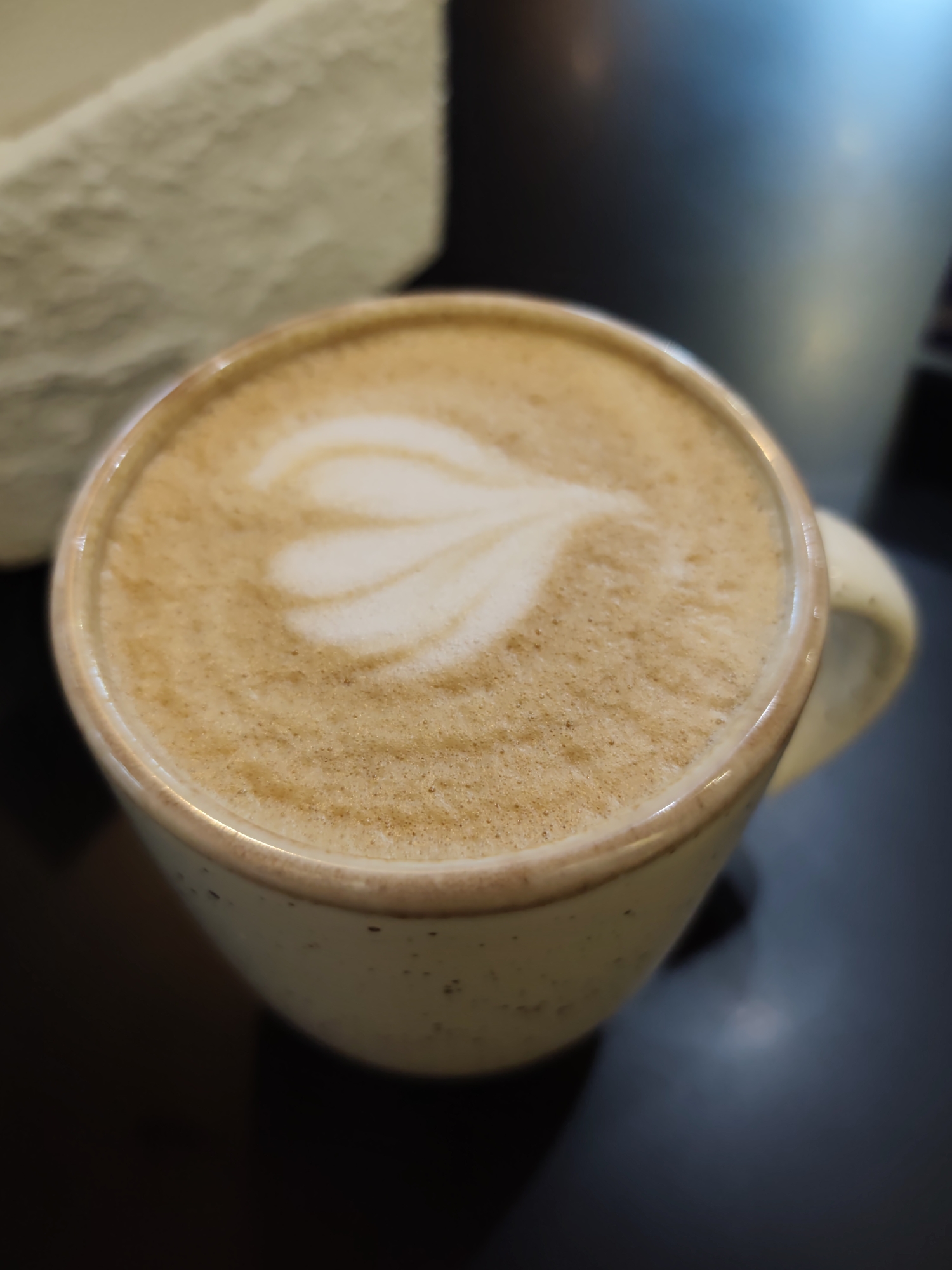 A close-up, eye-level, slightly overhead shot captures a latte art design of three interconnected hearts in a speckled, light-colored ceramic mug. The mug, with a light brown rim, sits on a dark, reflective surface, likely a table. The background is blurred, showing a pale, textured object on the left and a darker, reflective surface with bluish highlights on the right. No people are visible, and no specific location in Sochi, Russia, can be identified from the image.