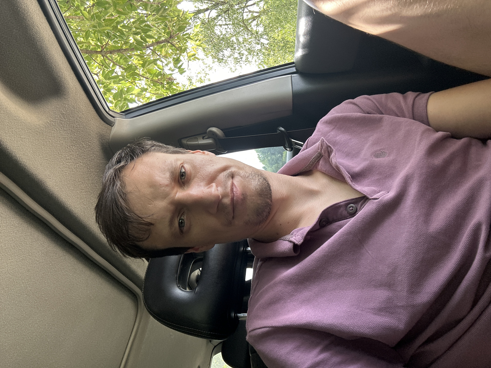 A man is seated inside a car, with his head and upper torso visible. He is wearing a light purple polo shirt. The car's interior includes a headrest and a sunroof. Through the open window on the right side of the frame, lush green trees and foliage are visible, suggesting an outdoor, possibly rural or suburban setting. The lighting indicates it is daytime, with natural sunlight illuminating the scene. The specific location in Makiv, Ukraine, is not discernible from the visual information provided.